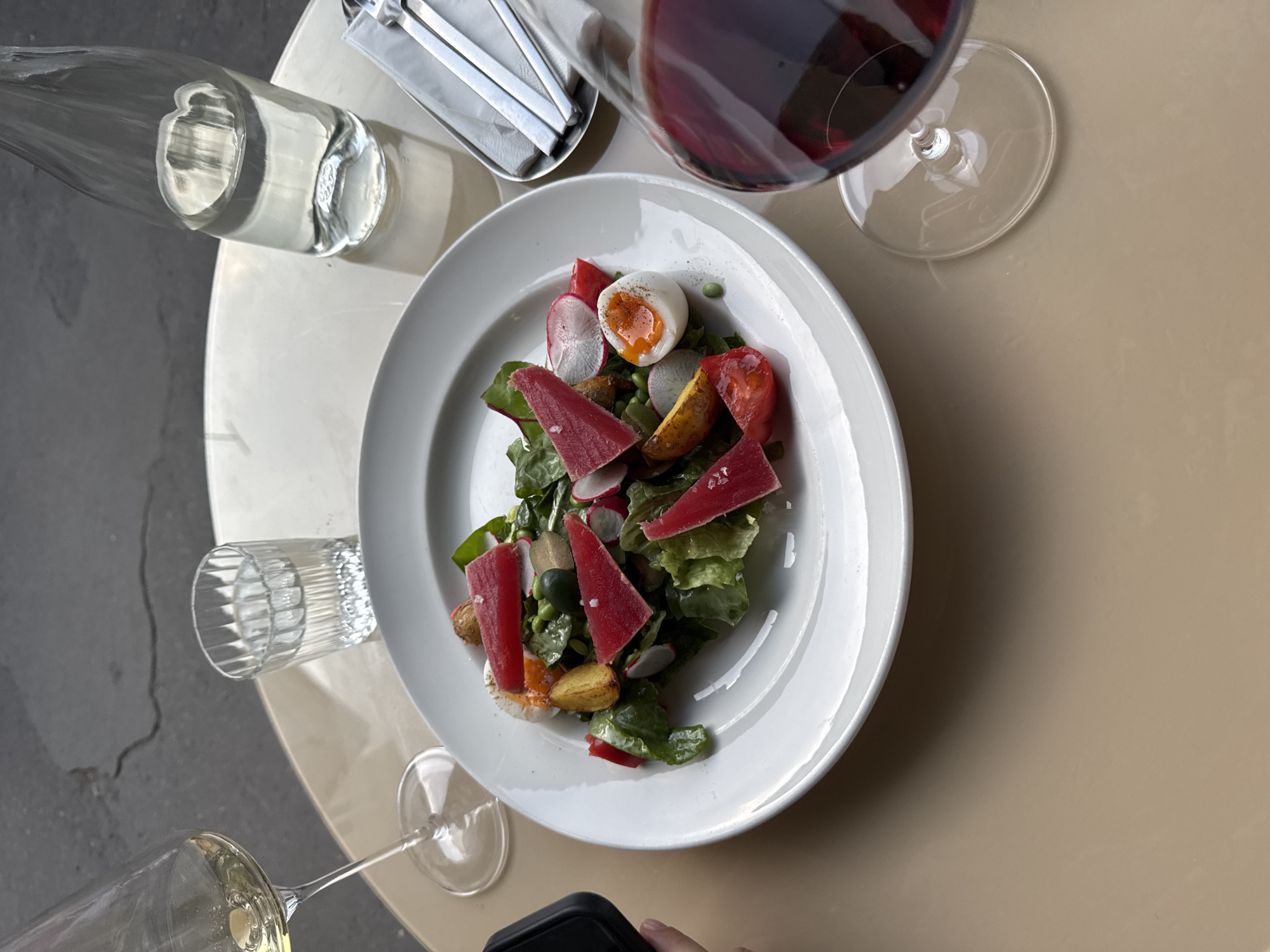 The image shows a table setting, possibly in a restaurant. On a round table are a bottle of water, a holder with cutlery, a glass of red wine, and a white plate containing a salad. The salad includes lettuce, radishes, tomatoes, boiled eggs, and what appears to be seared tuna or another red meat. There are also a small glass of clear liquid and a glass of white wine in the bottom corner of the image, as well as a phone. The table is situated next to what appears to be a concrete surface. The image location in the city of Moscow, Russia, cannot be verified from the image.