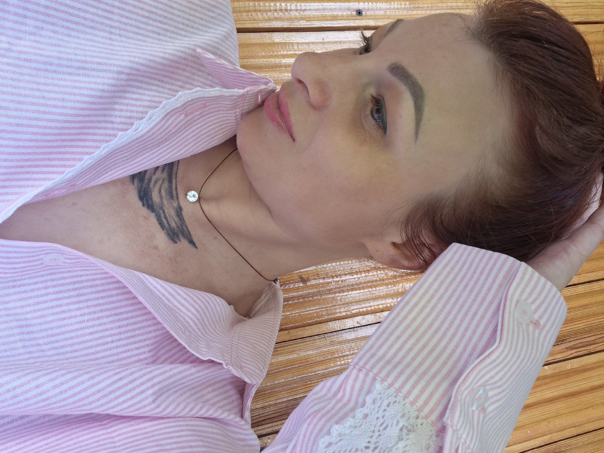 A woman with auburn hair, pulled back, is shown from the chest up, looking down and to her left. She is wearing a pink and white striped button-up shirt with lace trim on the cuffs. A delicate necklace with a single, clear pendant hangs around her neck, resting just above a tattoo on her chest. The background appears to be a wooden wall, possibly an exterior or interior wall of a rustic structure. The lighting suggests an indoor setting with natural light. No specific location in Забалоцкі сельскі Савет, Belarus can be determined from the image alone.