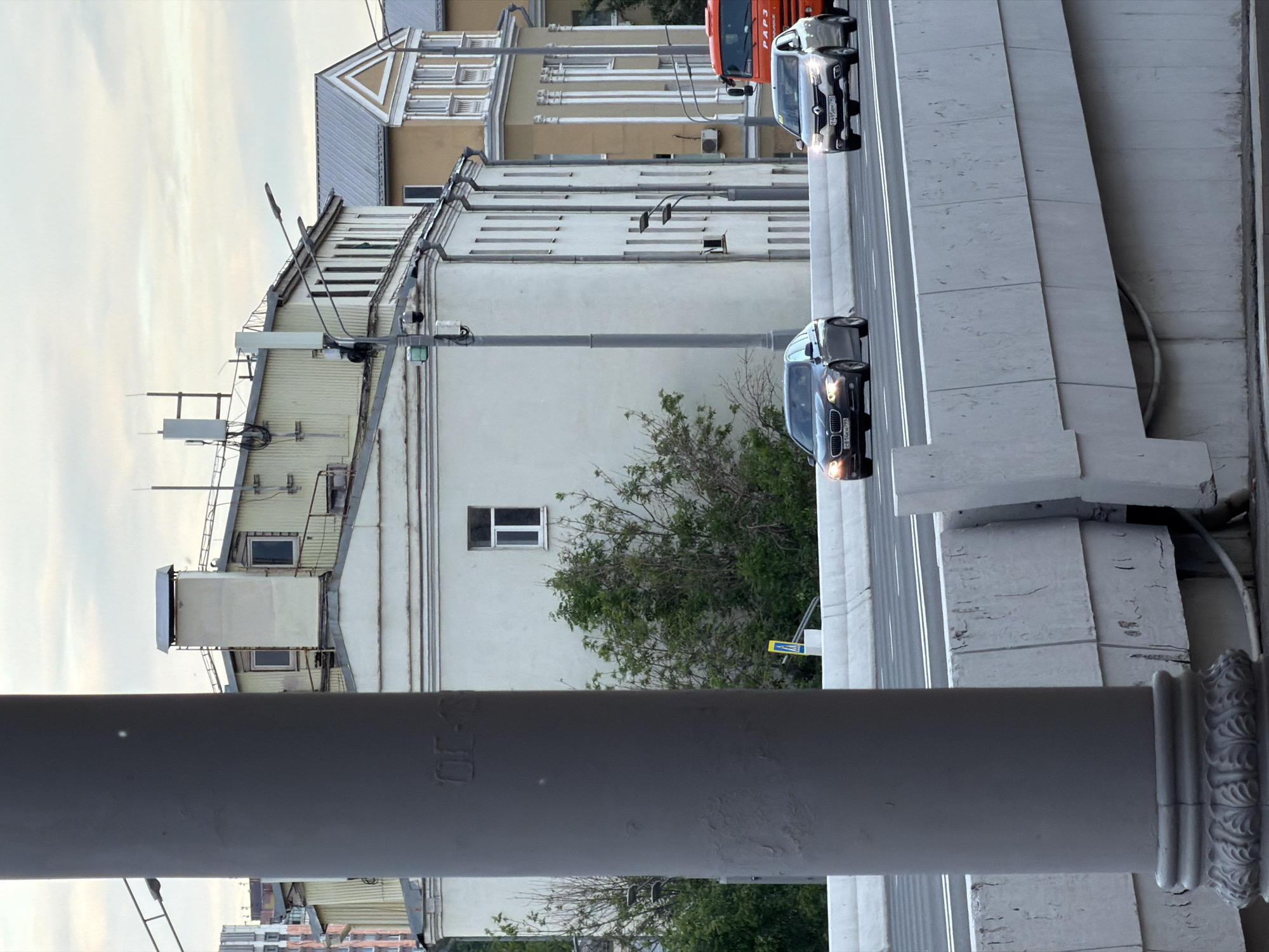 The image shows a vertical view of a street in an urban environment, likely in a European city. A raised section of road or bridge runs horizontally across the upper portion of the frame. Two vehicles, a dark sedan, and a Renault van, are on the roadway. Buildings are visible in the background, including some with older architectural styles. There is a gray concrete column and bridge support in the foreground on the lower right. The sky is overcast. The overall impression is one of urban infrastructure.