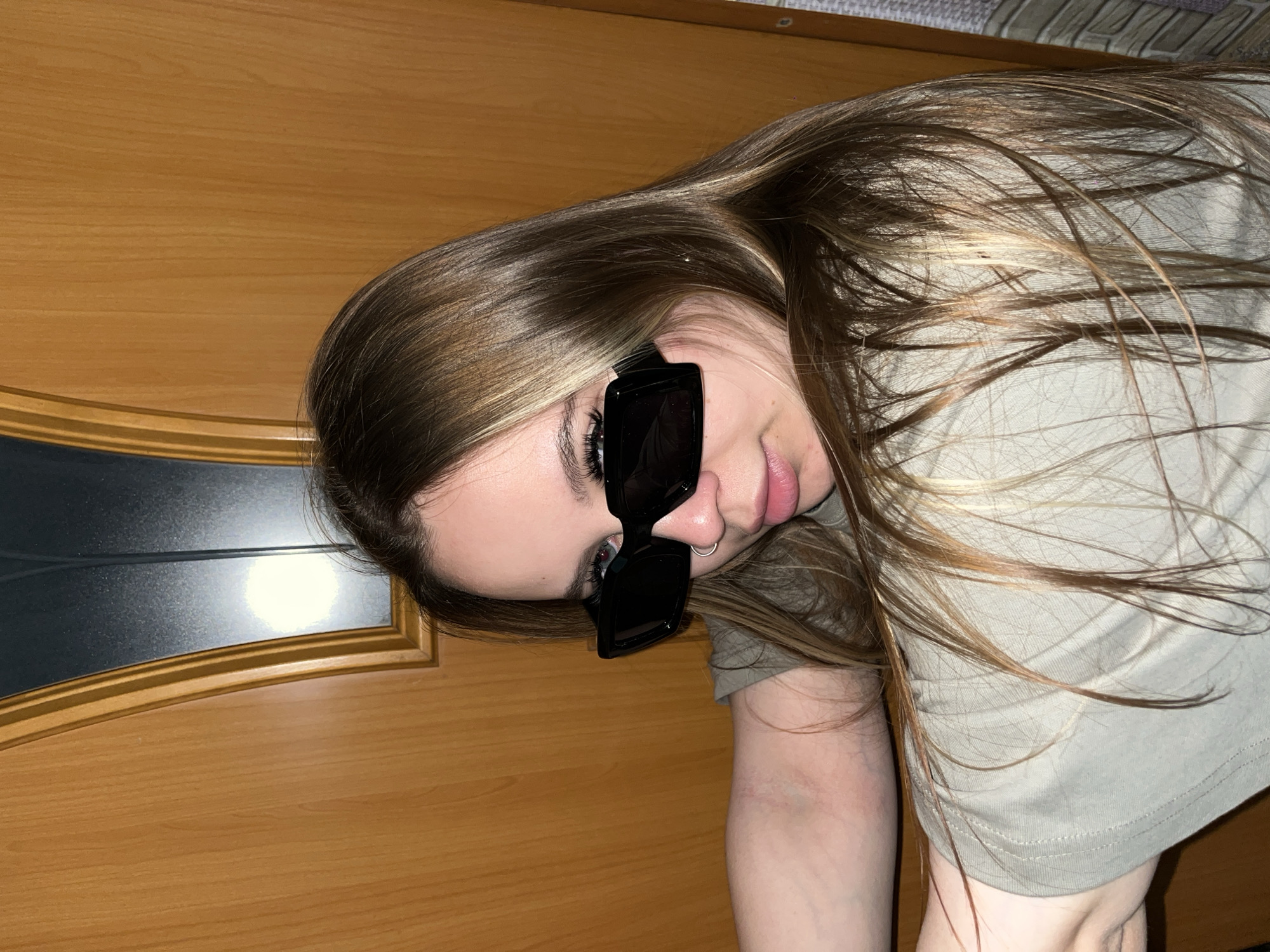 An eye-level shot captures a young person, likely female, with long, light brown hair streaked with lighter highlights, lying in a relaxed, casual pose. Her head is tilted, and she is looking towards the viewer. She is wearing black, rectangular sunglasses that cover most of her eyes, though glimpses of light-colored irises and long, dark eyelashes are visible underneath. A small silver nose ring is prominent on her nostril. Her lips are slightly parted.
She is dressed in a plain, light olive green t-shirt. Her hair is spread out around her head, resting on what appears to be a light-colored, possibly patterned fabric, likely bedding or a textured wall covering.
The background primarily features warm-toned wooden furniture, which appears to be a bed frame or headboard. On the left, a section of the wooden surface has a dark, reflective panel or mirror inserted, framed by the wood. The lighting suggests an indoor setting, possibly with artificial light, highlighting the hair and facial features. There is no visible text in the image. The scene is located in Port-Katon, Russia.
