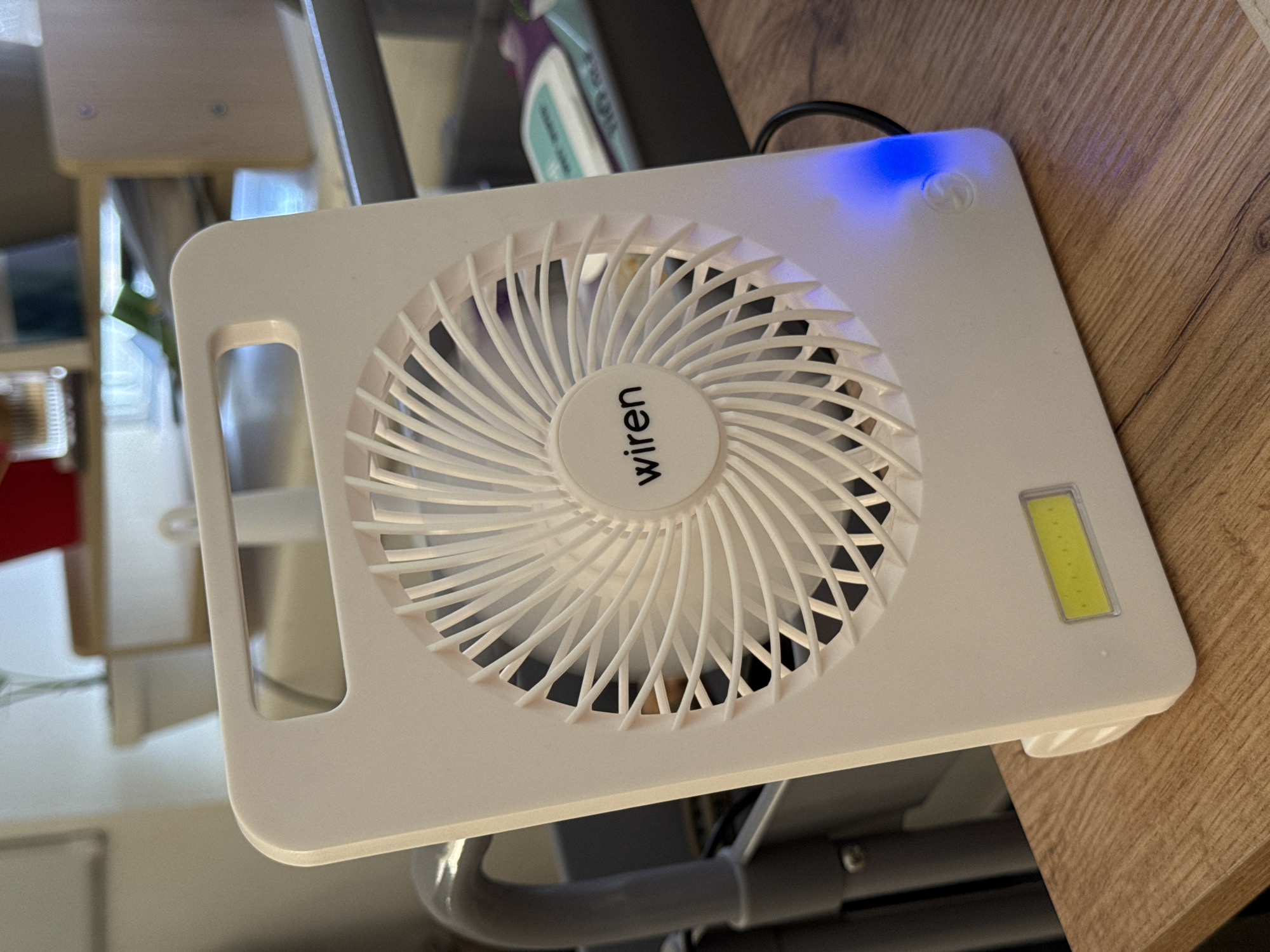 A white portable fan with a carrying handle is placed on a wooden surface. The fan has a circular grill with white blades and the brand name "wiren" is visible in the center. A rectangular yellow LED light is located on the front of the fan, below the grill. A blue light is illuminated on the bottom right corner of the fan, indicating it is on or charging. The background is blurred, showing a desk with shelves and some items on them. The scene is indoors, in what appears to be an office or a study environment.
