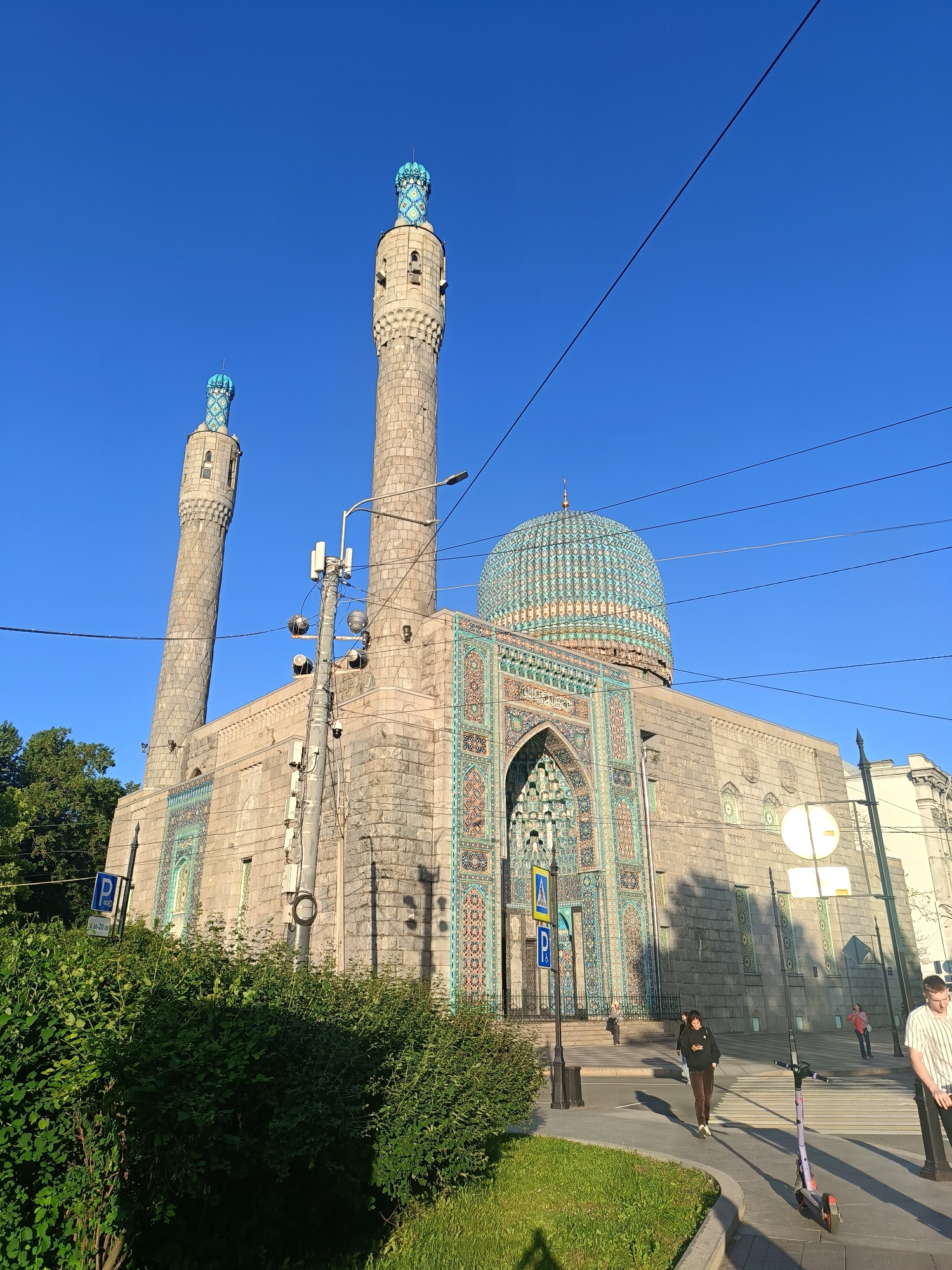 The image shows the Saint Petersburg Mosque, a prominent landmark in the city of Saint Petersburg, Russia. The mosque is an impressive structure with a large blue-tiled dome and two tall minarets, also adorned with blue tiles. The facade is decorated with intricate geometric patterns and Arabic calligraphy. In the foreground, there is a street with a sidewalk, a grassy area with bushes, and a few people walking. There is also a parked electric scooter. Overhead, power lines crisscross the clear blue sky. The lighting suggests it is a sunny day.