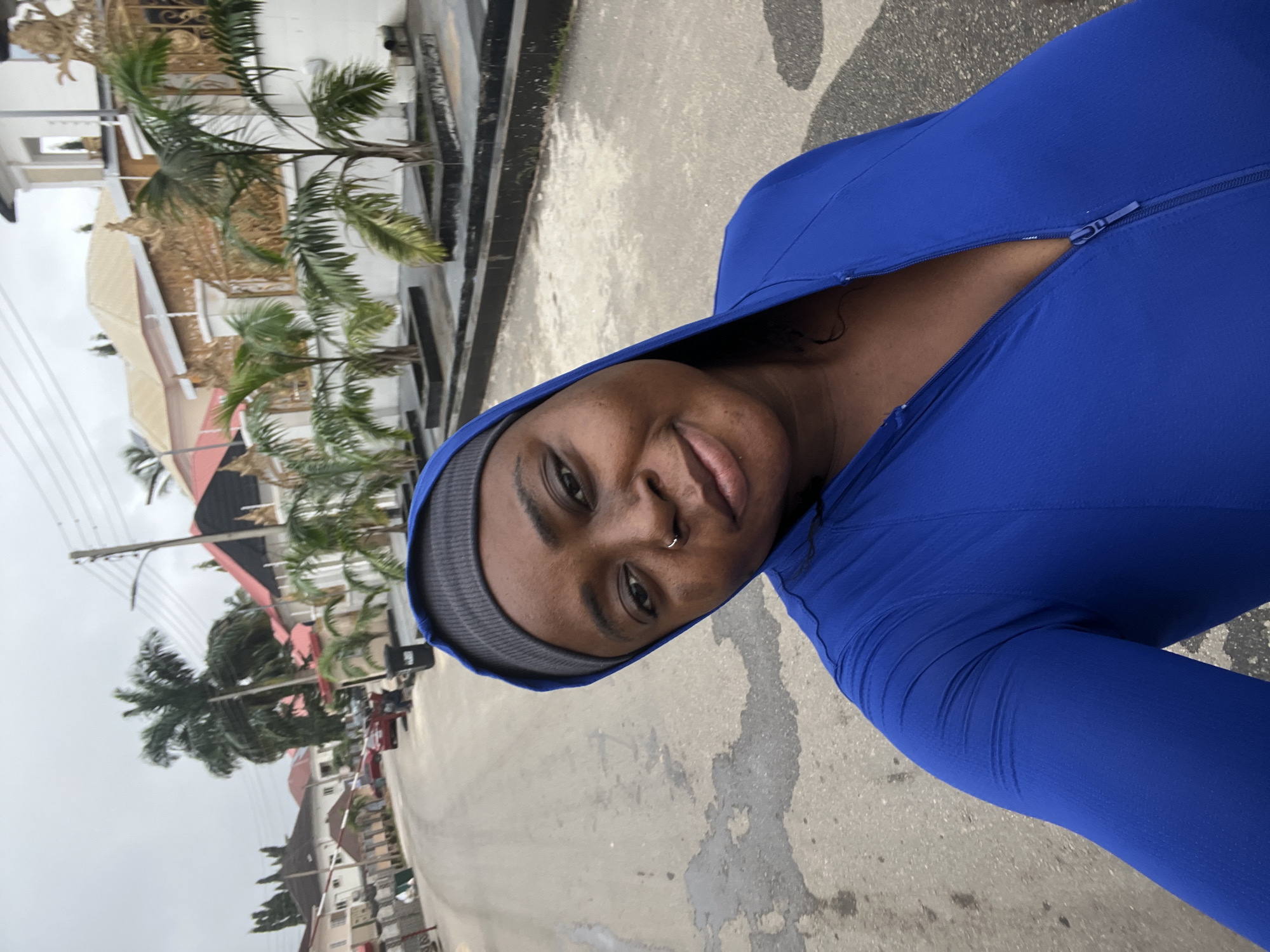 A woman with a nose ring is captured in a selfie taken outdoors on a cloudy day in Abuja, Nigeria. She is wearing a blue hooded jacket with a zipper partially open, revealing her décolletage, and a grey beanie. The background shows a paved street lined with modern houses and palm trees, characteristic of Abuja's residential areas. Several cars are parked along the street, and a trash bin is visible near the curb. The architecture of the houses suggests affluence, with ornate gates and decorative fencing. The sky is overcast, indicating a potential for rain.