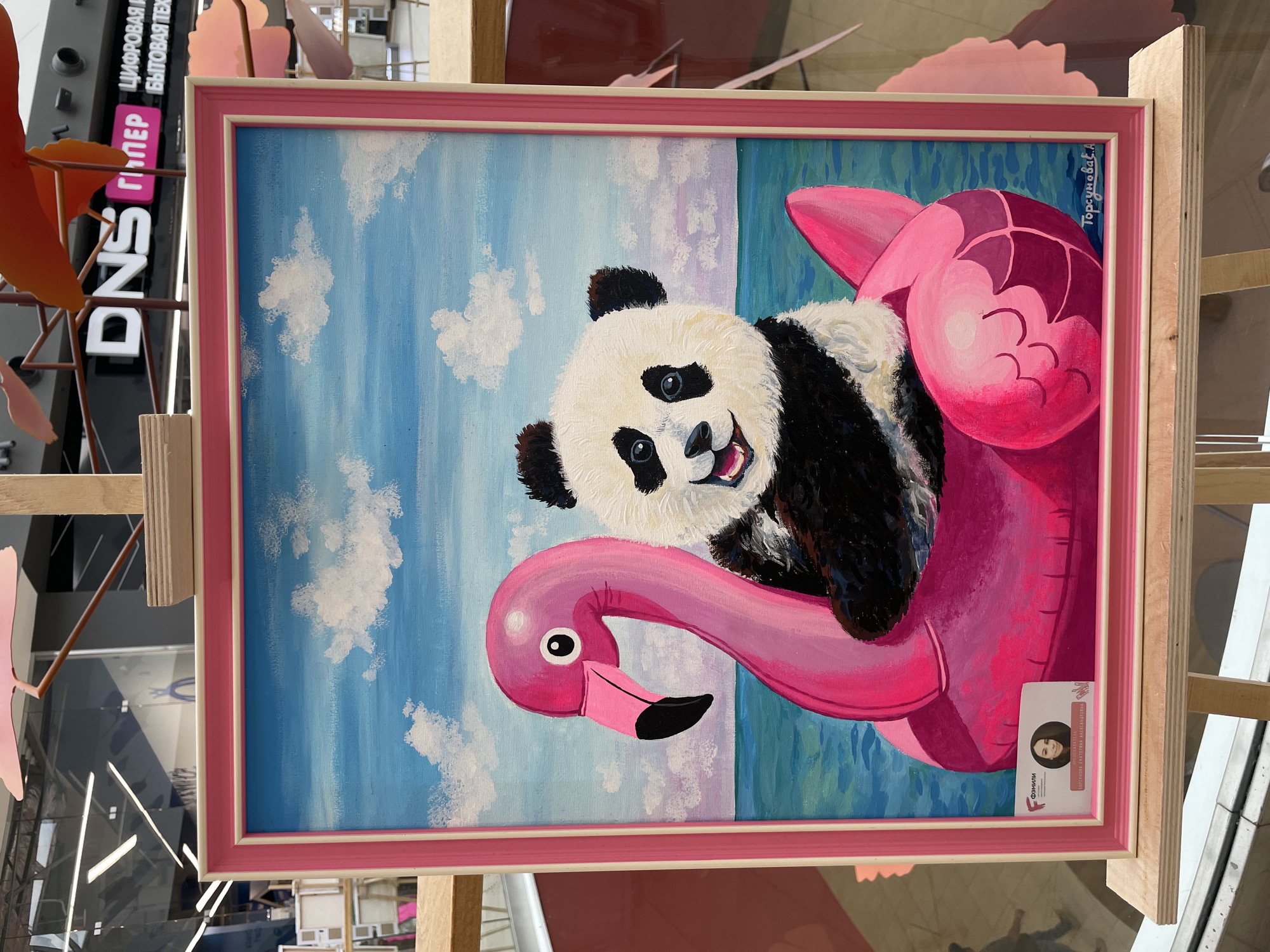 This media file shows a painting of a panda riding a pink inflatable flamingo. The panda is depicted with a happy expression, its head tilted and mouth open. The flamingo is stylized with bright pinks and purples, floating on a blue and green body of water under a bright blue sky with fluffy white clouds. The painting is framed with a pink and white border.
A small sticker is attached to the bottom right corner of the frame, featuring a portrait of a young woman and text in Russian. The text on the sticker appears to be an artist's name and perhaps information about the artwork. The text "Торгунова Е.А." is visible in the top right corner of the painting itself, indicating the artist's name.
The painting is displayed on an easel, and in the background, there are hints of an indoor environment, possibly a gallery or exhibition space. A sign with large white letters "DNS" is visible to the left of the painting, along with other signs in Russian text. The lighting suggests it is daytime. The overall mood of the painting is cheerful and whimsical. The location context provided is Perm, Russia.