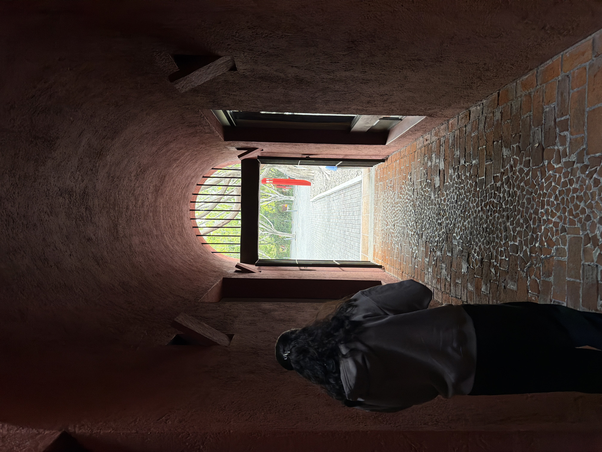 The image shows a person standing in what appears to be the Camelia Pasaje, a building in the city of Puebla, Mexico. The interior space has a rounded wall made of a rough, red-toned material. A window provides an outside view of a building, foliage, and a glimpse of what might be a basketball hoop. The other wall, on the right side of the image, is constructed of reddish-brown brickwork. Wooden slats are seen on the rounded wall. The person stands in the darkness of the space, facing towards the window. The person is wearing a white jacket or shirt and black pants. Their dark hair is visible.