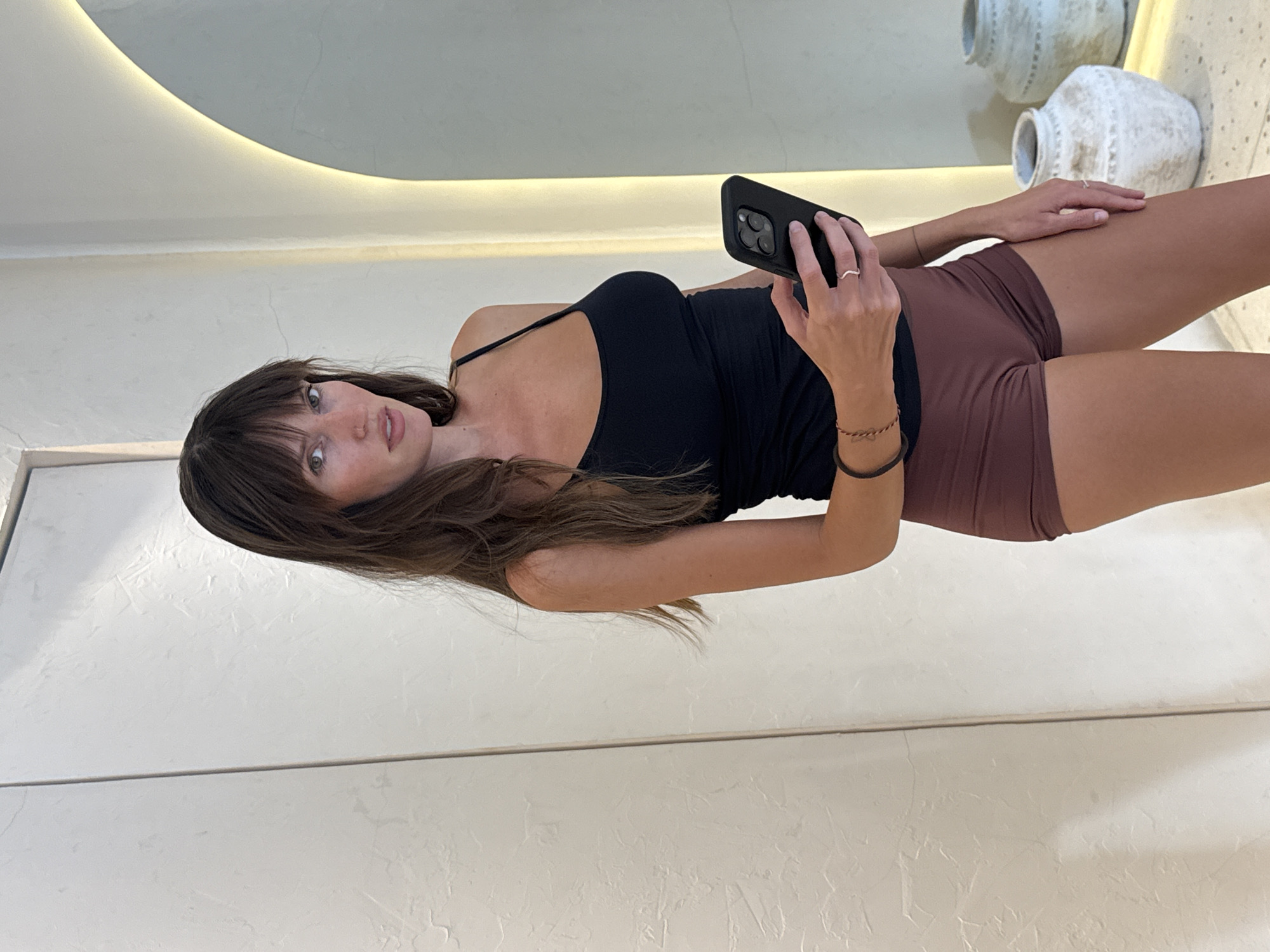 A woman with brown hair and bangs is holding a black smartphone and taking a photo of herself in a mirror. She is wearing a black tank top and maroon shorts. Behind her, partially visible in the mirror, are two decorative white vases. The location is likely Cemagi, Indonesia. The woman appears to be indoors.
