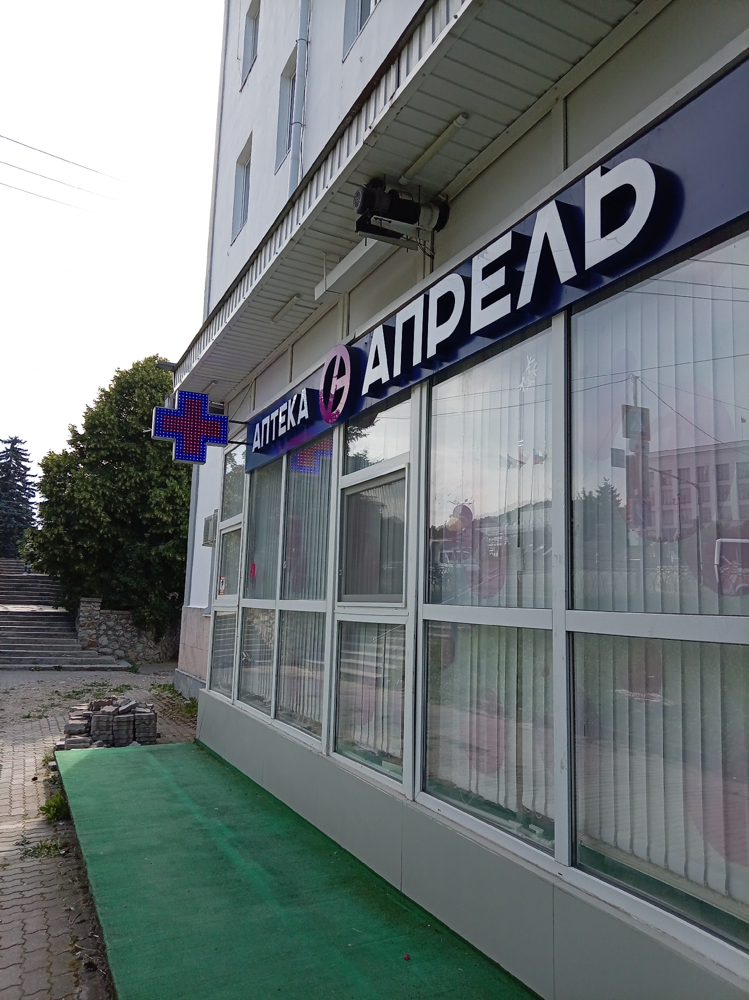 The image shows the facade of a pharmacy building with a prominent sign that reads "АПТЕКА АПРЕЛЬ" in large white letters against a dark blue background. To the left of the sign, a blue and red illuminated cross, a common symbol for pharmacies, is visible. The building has large windows with blinds, reflecting the environment outside. In front of the building, there is a green carpeted area, and to the left, a set of concrete stairs leads up a slight incline, flanked by trees. A stack of paving stones is visible on the left side of the image, near the stairs. The sky is overcast. The scene appears to be an exterior view of a commercial establishment in a city setting. The pharmacy is located on Kalinin Street in Zheleznovodsk, Russia.
