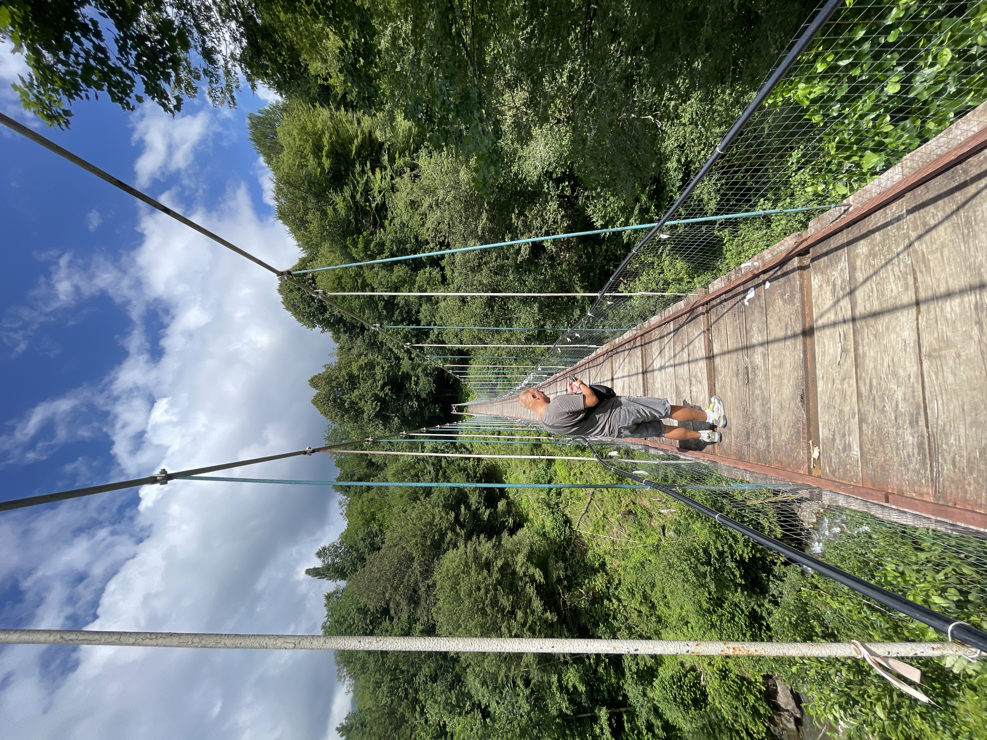 In Tqwarchal, Abkhazia, a man stands on a wooden suspension bridge, looking down at his phone. He is wearing a grey t-shirt and grey shorts. The bridge is surrounded by lush green trees and has a mesh railing. The sky is blue with scattered white clouds. A river can be seen flowing below the bridge. The bridge appears to be aged, with weathered wooden planks and metal supports. The scene captures a moment of pause amidst the natural beauty of the Abkhazian landscape.