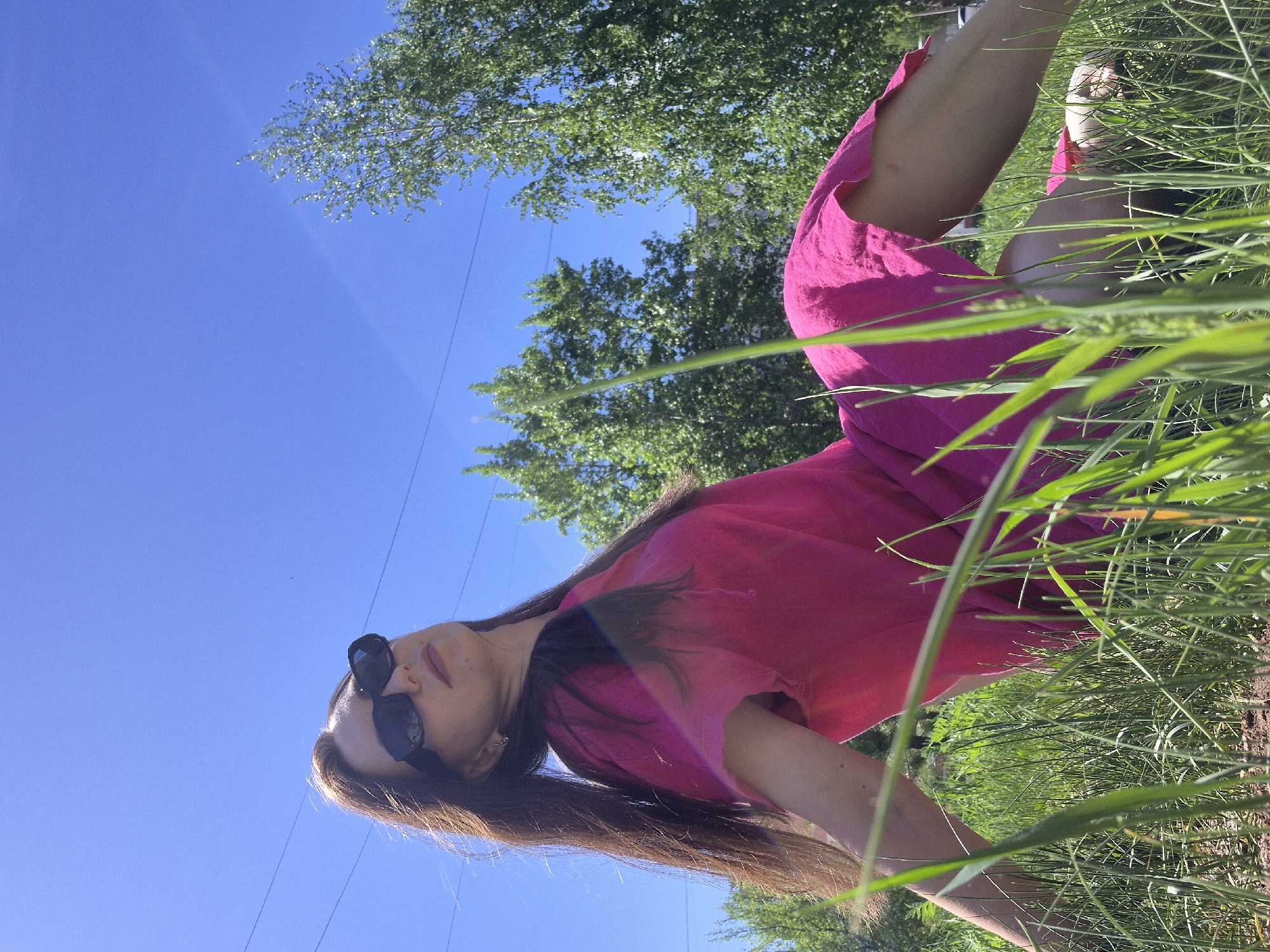A low-angle shot depicts a woman wearing a pink dress and sunglasses. She is situated amongst tall grass, with trees and a clear blue sky visible in the background. The location is unknown.