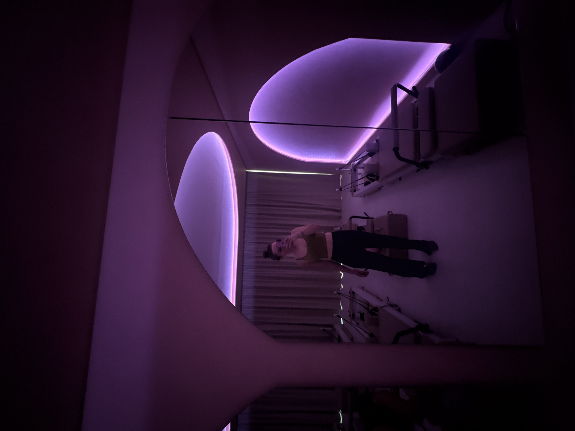 The image shows a woman in a dimly lit room with purple lighting, possibly a fitness studio. The studio is located in Mexico City, Mexico. The woman is wearing a crop top and dark pants. Several Pilates reformer machines are visible in the room. The lighting creates a modern and somewhat futuristic aesthetic.