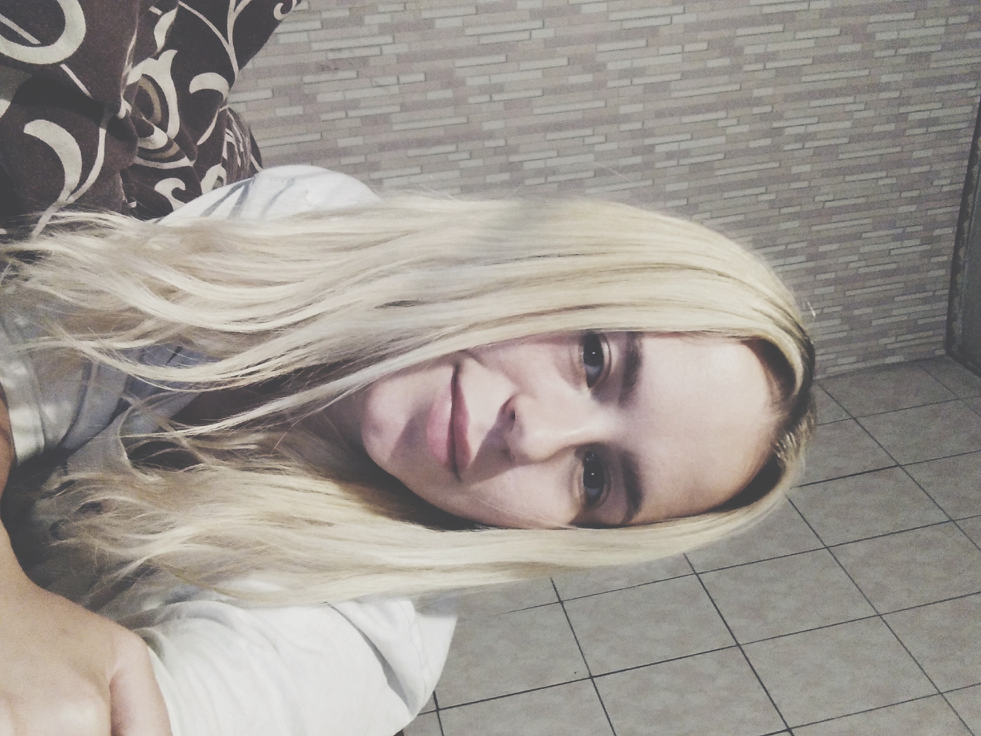 An overhead medium shot shows a young woman with long blonde hair, facing forward and looking at the camera. She is wearing a white shirt and is partially covered by a blanket with a brown and white pattern. The background consists of a textured wall with a vertical striped pattern on the left and a tiled wall on the right. The scene appears to be indoors, possibly in a bedroom or living area.
