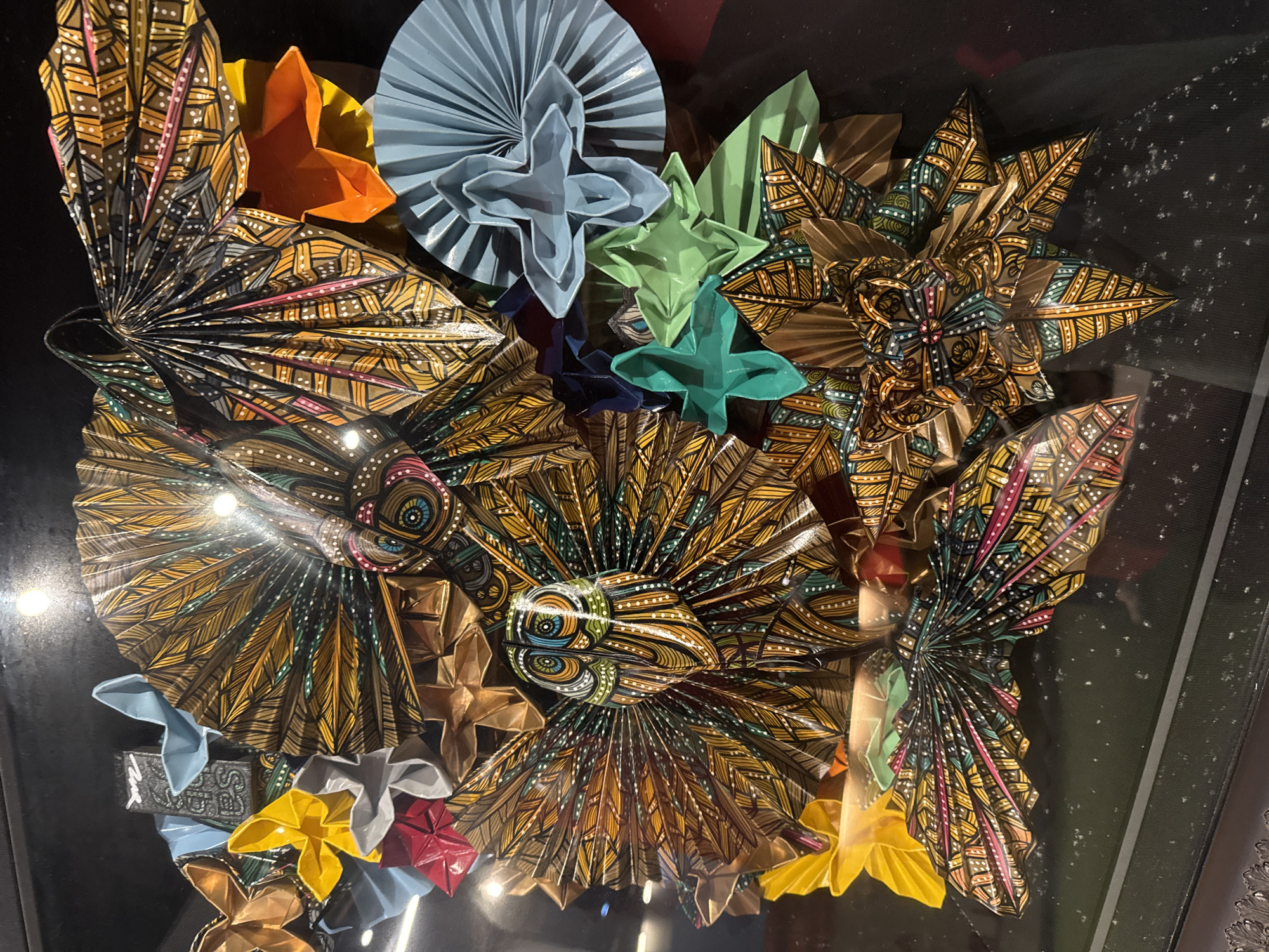 The image shows an intricate display of colorful origami figures, predominantly shaped like flowers and birds, arranged against a dark background. The origami pieces feature vibrant patterns with geometric designs, feathers, and floral motifs in shades of gold, brown, green, blue, yellow, red, and pink. One prominent origami figure resembles an owl with a detailed face, surrounded by fan-like wing structures. Other origami shapes include layered petals, starbursts, and folded paper fans. The overall presentation is artful and appears to be a decorative arrangement. There is no discernible information about the city of Kuta, Indonesia, or any specific location within it, in the provided image.