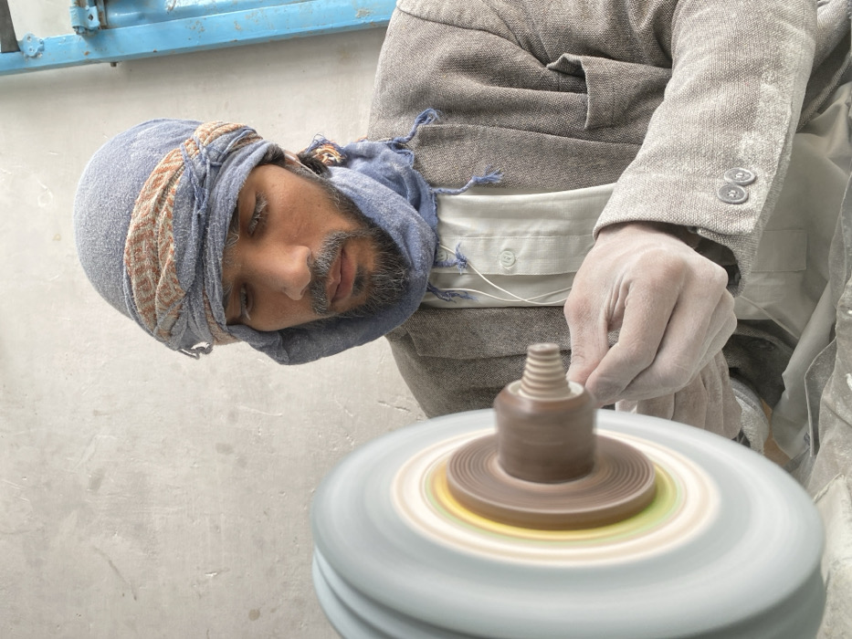 An adult male is captured in a close-up, working in a dusty workshop located in Kandahar, Afghanistan. He is positioned from the chest up, wearing a grey sport coat over a light shirt, and a blue head covering (turban/scarf) wrapped around his head and neck. His face, beard, and hands are visibly covered in white dust. He is focused intently on a small, light-colored, threaded wooden object, which he holds with both hands, also dusted white. This object is being worked on by a rapidly spinning machine component, consisting of a large grey and white wheel with concentric brown, yellow, and green rings near its center, attached to a brown cylindrical spindle. The man is wearing white wired earbuds. The background features a plain, light grey textured wall on the left, and a partial view of a bright blue metallic structure on the right.