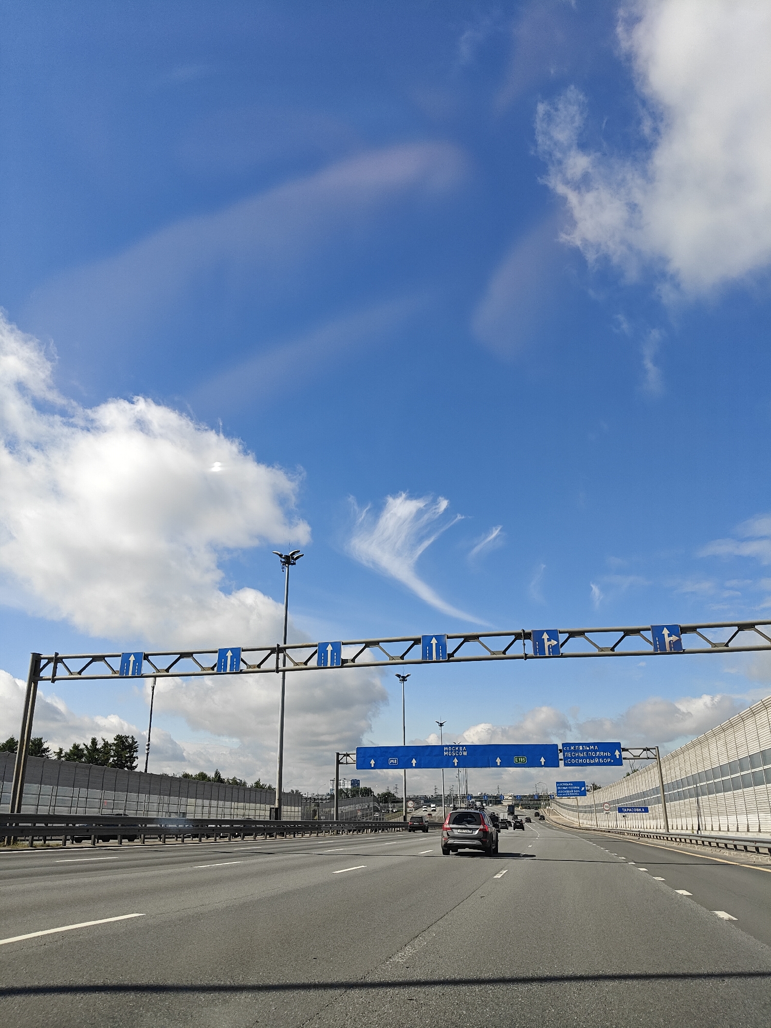 The image shows a view from a car driving on a multi-lane highway, likely in or near Pushkino, Russia, under a bright blue sky with scattered clouds. Traffic signs are visible overhead, indicating directions to Moscow and other destinations, including "Pesnie Polyani" and "Sosnovy Bor." Cars are traveling in the same direction as the viewer. The highway is flanked by sound barriers on the right and trees and buildings in the distance on the left. The overall scene depicts a typical highway environment on a clear day.
