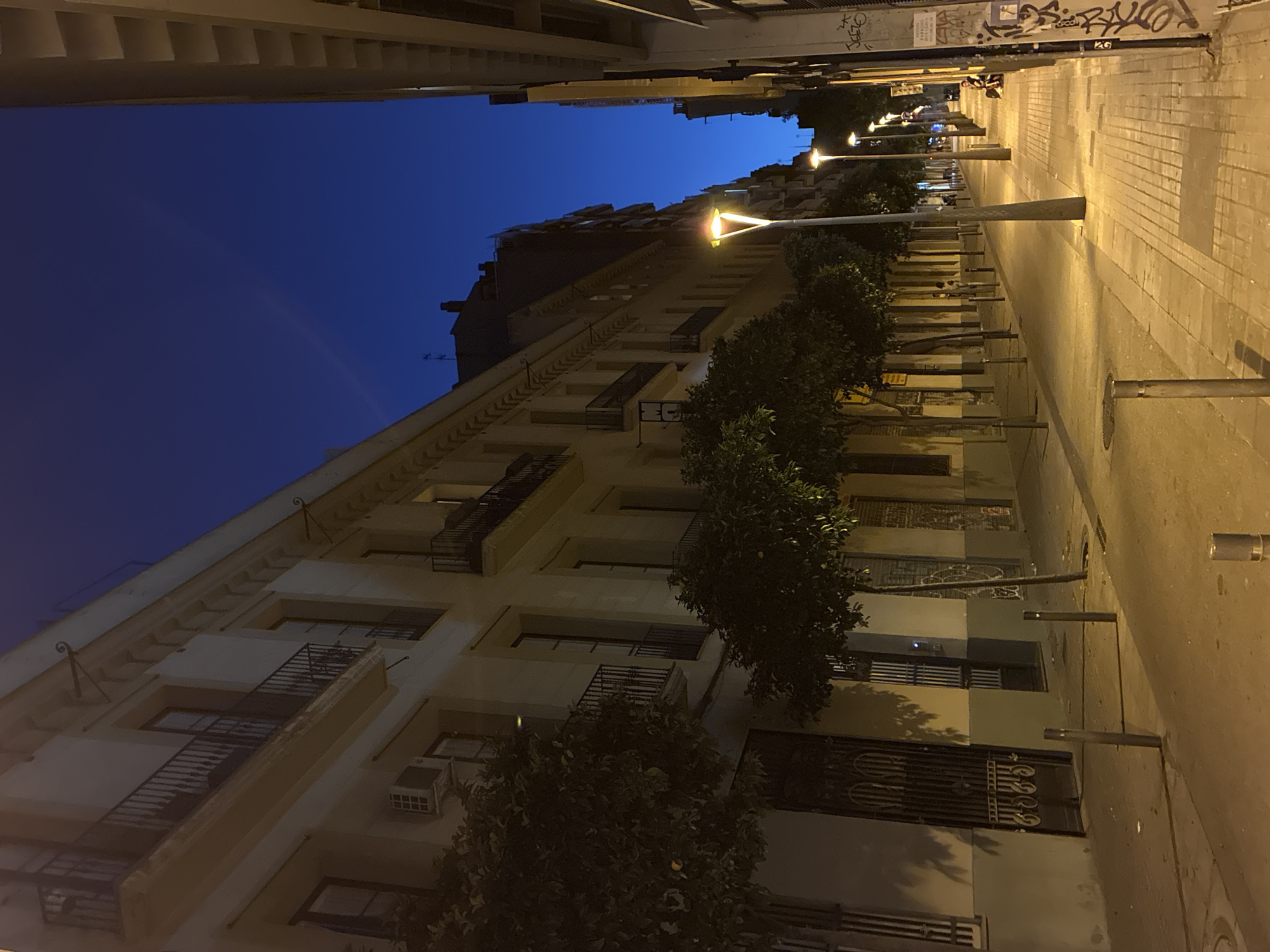 This image depicts a quiet urban street scene in Barcelona, Spain, captured at twilight or early night. The sky above is a deep, clear blue, suggesting the residual light of dusk.
On the left, a long row of multi-story buildings, characterized by light-colored (beige/off-white) facades and classical architectural details, stretches into the background. Each floor features numerous windows and balconies with black wrought iron railings. An air conditioning unit is mounted on the second story of the prominent building in the foreground. A series of small trees, identifiable as orange trees by their dark green foliage and visible small, round, orange fruits, line the sidewalk in front of these buildings. The ground level displays ornate, dark metal gates and barred windows.
The right side of the street features a wide sidewalk paved with light-colored square tiles. A procession of modern streetlights, each with a distinctive upward-pointing, V-shaped light fixture, emits a warm yellow glow that illuminates the path and recedes into the distance. Beyond the sidewalk, multi-story buildings with a darker facade line this side of the street. Graffiti is present on a pillar or wall section in the extreme foreground on the right. One person is visible on the far right sidewalk, seated or resting against a structure. Another faint figure is discernible on the left sidewalk further down the street.
The street itself is paved with asphalt, appearing dark, and features several short, grey metal bollards strategically placed along both sidewalks. Manhole covers are visible on the street surface. The overall scene is calm and well-lit by artificial light, indicative of an urban environment in Barcelona, Spain, given the characteristic architecture, ubiquitous orange trees, and modern street furniture.