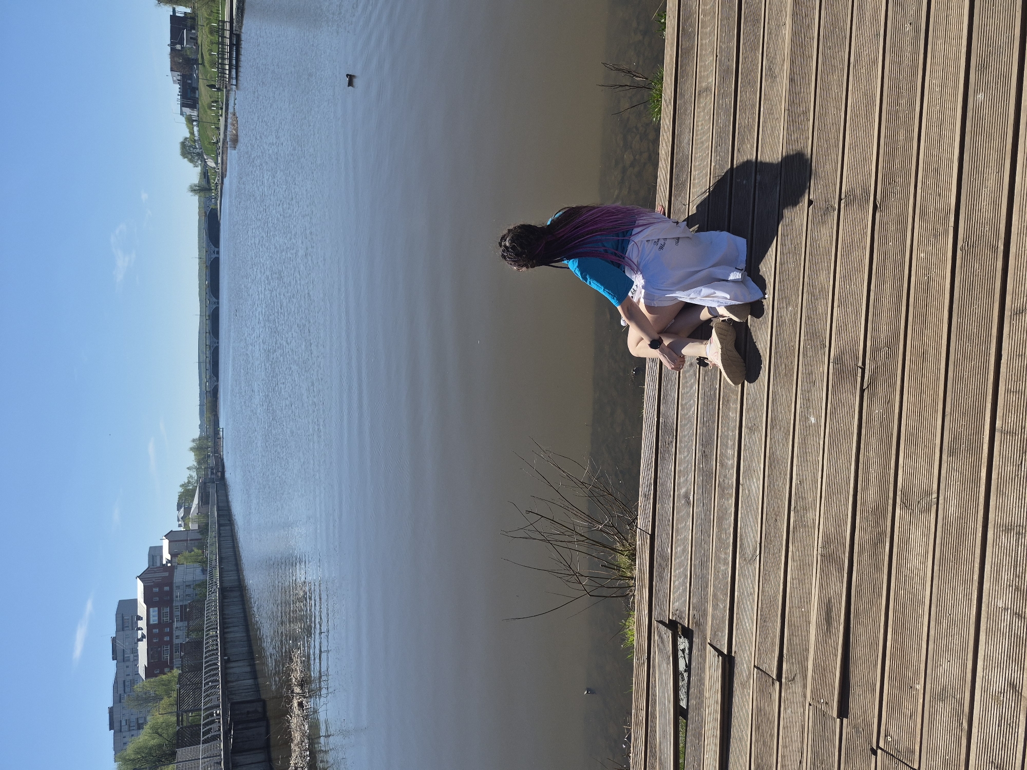 A long-shot captures a person sitting on a wooden boardwalk, looking out over a body of water, with buildings visible in the background. The boardwalk is made of parallel wooden planks. A person with dark hair streaked with pink is kneeling or sitting on the boardwalk, wearing a blue shirt and white shorts or skirt, facing the water. The water extends horizontally across much of the frame, with reflections and a duck visible on its surface. The buildings on the distant shore are mixed-use, multi-story structures. The sky is clear blue. The overall scene has a bright, sunny ambiance. This is in Votkinsk, Russia.