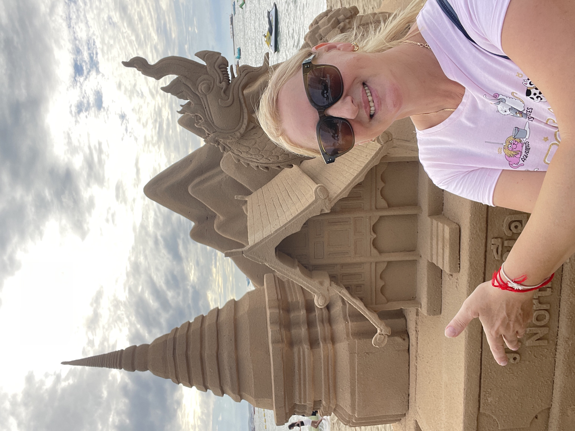 A blonde woman wearing sunglasses and a pink t-shirt with cartoon cat graphics poses for a selfie in front of a sand sculpture on a beach in Pattaya City, Thailand. The sand sculpture depicts a temple structure, a mythical creature resembling a dragon or serpent, and mountains in the background. The woman has a red and white bracelet on her wrist and a gold chain necklace. Her left hand is extended towards the sculpture. The sea and a jet ski are visible in the background under a cloudy sky. The word "North" is visible on the base of the sand sculpture.