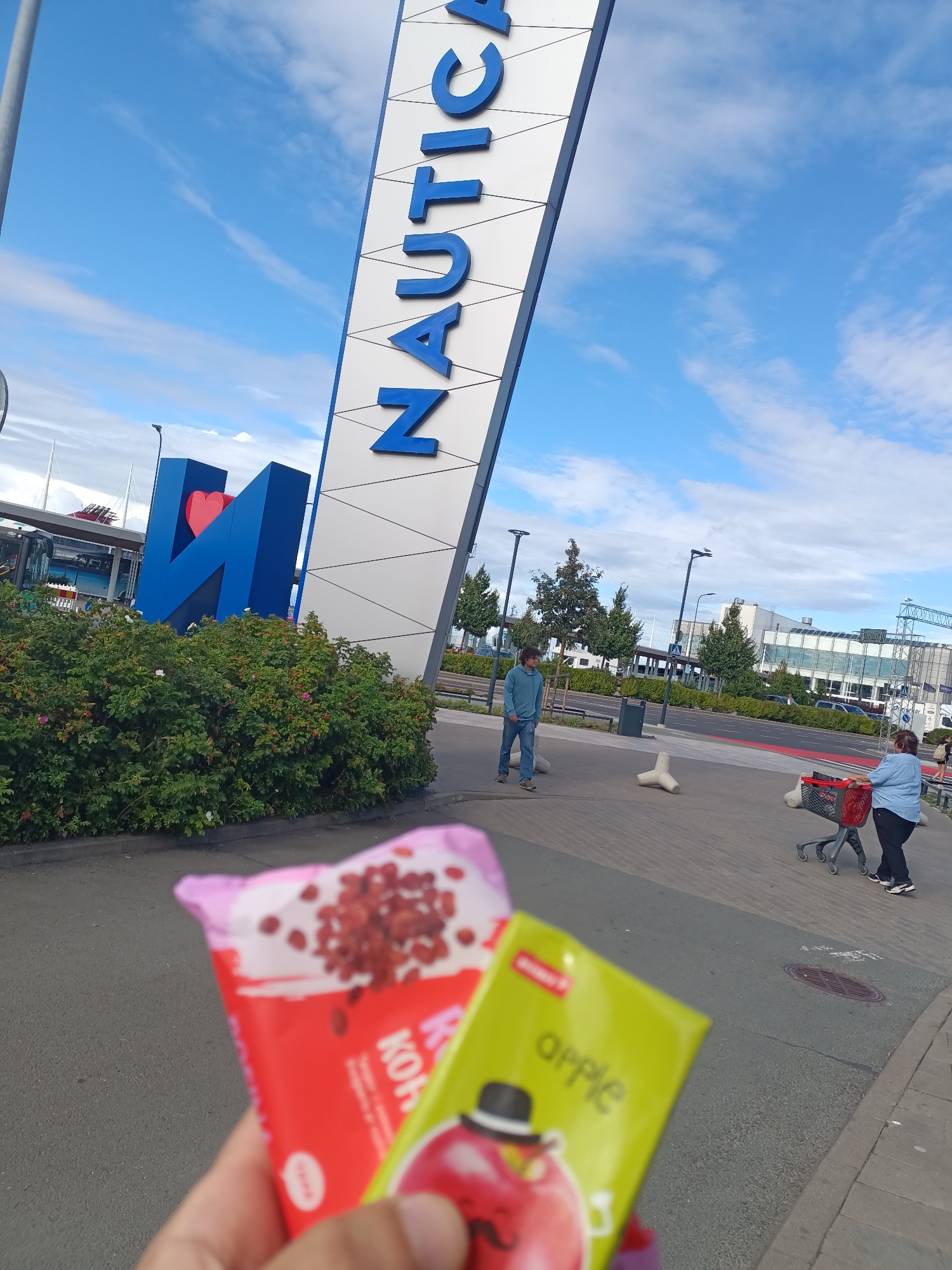 The image shows a close-up of two ice cream bars being held in the foreground. The ice cream bars are on the right side of the frame. In the background, there is a large blue sign with white diagonal lines and blue lettering that reads "NAUTIC". To the left of the "NAUTIC" sign, there is a large blue structure with a red heart inside. A person is walking towards the right of the frame, and another person is pushing a shopping cart. The scene appears to be in a public outdoor space, possibly a shopping center or a waterfront area. The sky is blue with some white clouds. This location is the Nautica shopping center in Tallinn, Estonia.