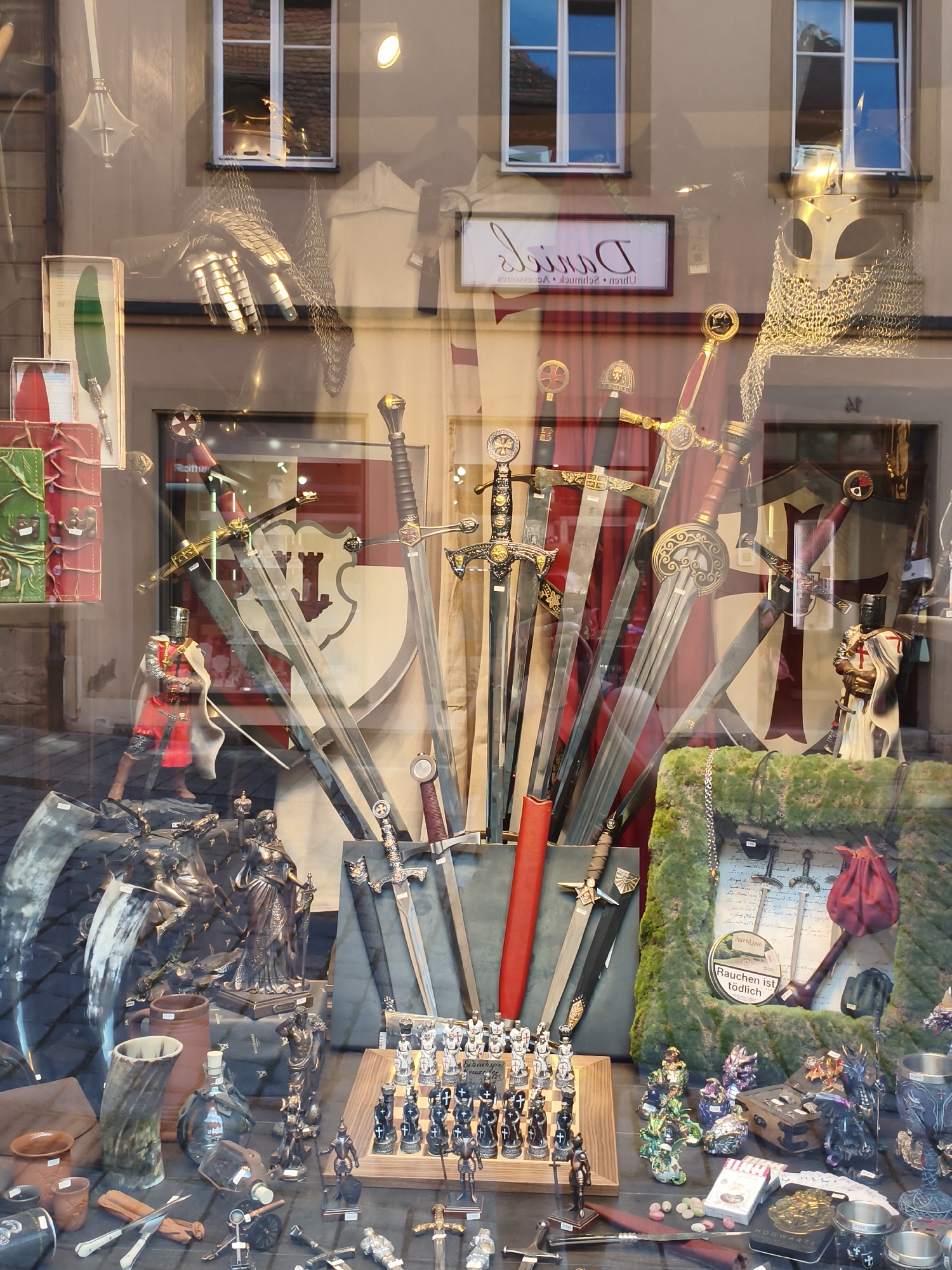 This image depicts a shop window display in Rothenburg ob der Tauber, Germany, featuring a collection of medieval-themed items. The window is filled with an array of swords, armor pieces, and decorative figurines, suggesting a historical or fantasy merchandise store.
Dominating the center of the display are several large swords with ornate hilts and blades. To the left, a knight figure in red armor stands with a cape, positioned near a large decorative horn. Further to the left, a green quill and a red leather item are visible.
The background shows a reflection of a building with windows, indicating the display is set up behind glass. One of the reflections reveals two windows with tiled roofs visible inside. A sign with the name "Daniel's" is also reflected.
The lower part of the display showcases a chess set with medieval-themed pieces, miniature knight figurines, dragon figurines, and other decorative items. A framed display with small swords and a pipe is also visible on the right side of the window. The overall scene is a curated presentation of items related to knights, battles, and historical fantasy.