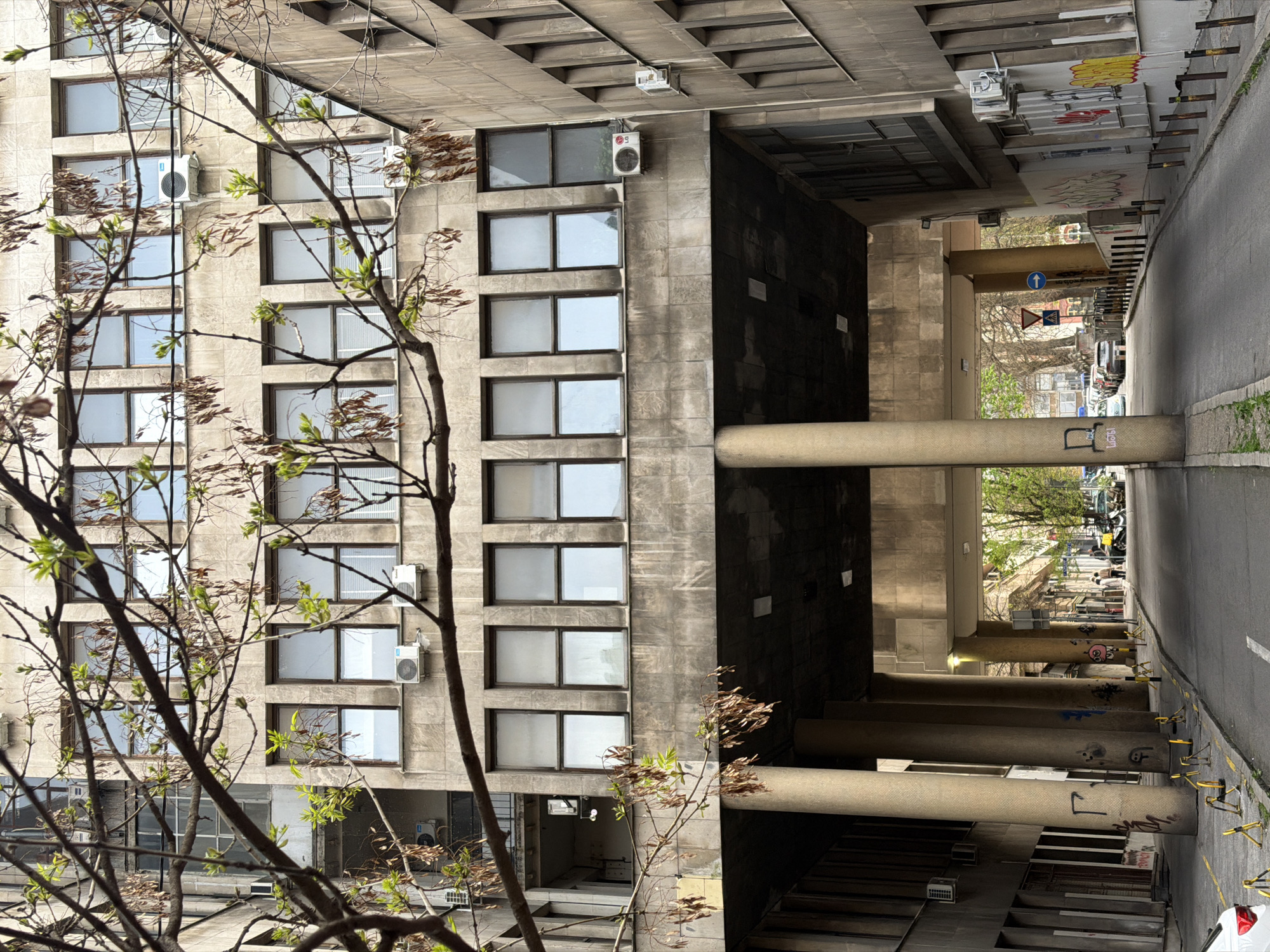 The image shows a building with rows of windows and air conditioning units. A bare tree with few leaves partially obscures the view of the building. The building appears to be part of a larger structure with an underpass or covered area supported by large concrete pillars. Graffiti is visible on the pillars. Beyond the underpass, a street scene is visible, with parked cars and pedestrians. The location is likely in Belgrade, Serbia, but confirmation would require local knowledge. The overall scene suggests a somewhat urban, possibly Brutalist architectural environment.