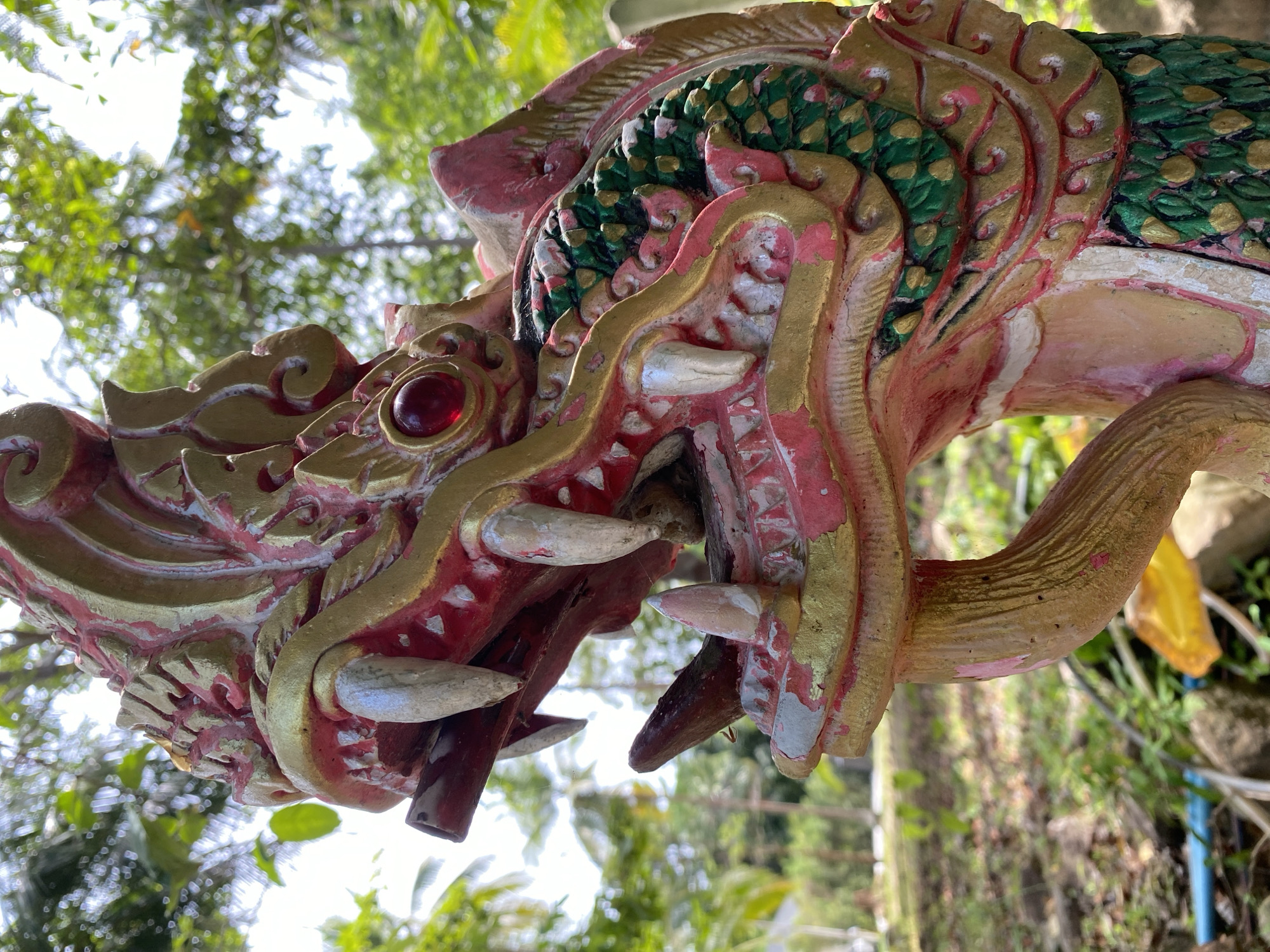 This is a close-up, eye-level, outdoor, vertical shot of a weathered, decorative statue of a mythical serpent-like creature, likely a Naga, in Koh Phangan, Thailand. The statue is predominantly red and gold with green scales visible on its neck. It features prominent, sharp, white teeth, a red jewel eye, and intricate decorative carvings. The background is blurred, showing lush green foliage, palm trees, and a bright sky, suggesting a tropical environment. The statue is positioned in an outdoor area with visible rocks and some scattered debris on the ground. The focus is sharp on the statue's head and neck, with a shallow depth of field blurring the surroundings.