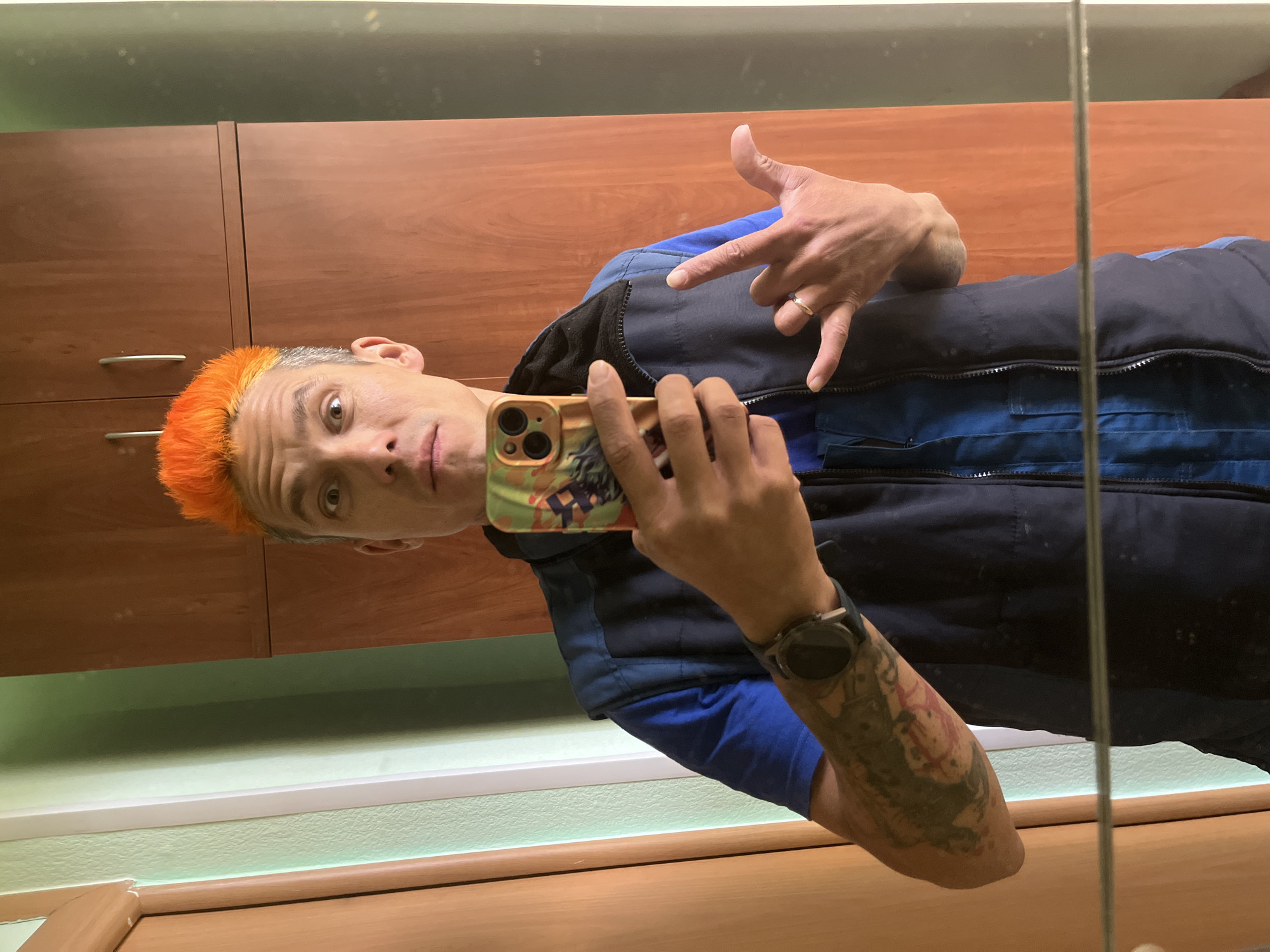 A man is taking a selfie in what appears to be a bathroom. He has orange hair and a tattoo on his forearm. He is wearing a blue shirt and a navy vest. He is holding a smartphone with a colorful case. He is making a hand gesture. The background includes wooden cabinets and light green walls. The location is unknown.