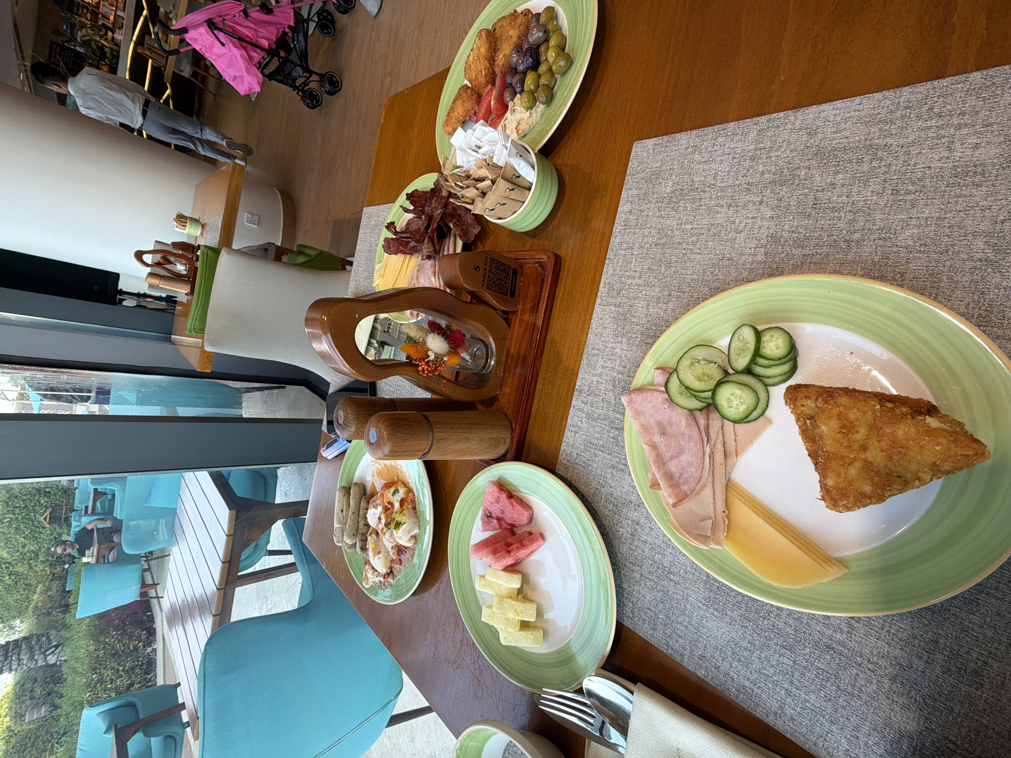 The image depicts an outdoor dining scene at a restaurant. The restaurant is likely situated in Dubai, United Arab Emirates, given the contemporary architecture and lush greenery visible through the large windows, suggesting a resort or a modern establishment.
In the foreground, a wooden table is laid with several plates of food. One plate contains a breakfast spread with sausages, eggs, and some red sauce. Another plate features cheese cubes and watermelon slices. A third plate, closer to the viewer, showcases sliced ham, cheese, cucumber slices, and a fried hash brown. Cutlery wrapped in a beige napkin is positioned to the left of the plates.
On the table, there are also salt and pepper grinders made of wood, a small decorative item with dried flowers in a glass, and a container filled with sugar packets. To the right, a green bowl holds more sugar packets. Further to the right, another plate holds fried chicken pieces, olives, tomatoes, and some kind of dip.
In the background, through large windows, a man is seen sitting at a table in an outdoor seating area. Beyond him, there are turquoise-colored chairs and tables arranged amidst greenery, indicating an outdoor dining or lounge area. In the far right background, a pink stroller is visible. A person in a white shirt and grey pants is walking towards the right side of the frame in the middle ground. The flooring appears to be tiled or wooden. The overall ambiance suggests a relaxed breakfast or brunch setting in a well-appointed venue.