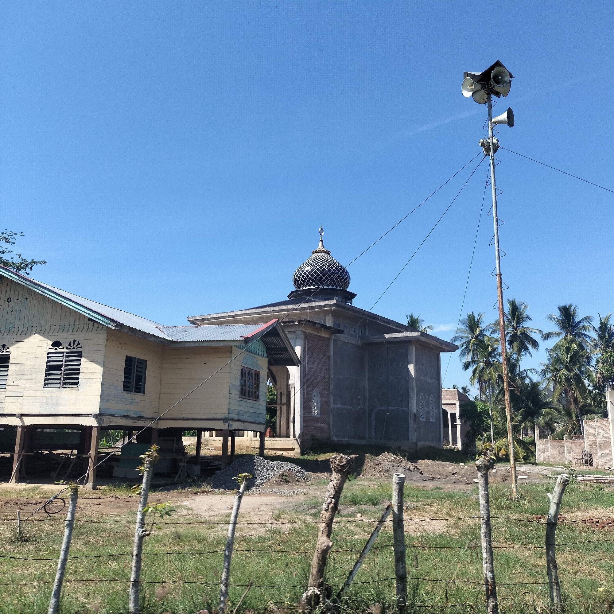A daylight exterior shot captures a tranquil rural scene in Tanjongkeumala, Indonesia, featuring a mosque, a traditional stilt house, and a prominent loudspeaker tower against a clear blue sky.
In the center-right of the frame, a mosque with concrete and brick walls is visible, topped by a distinctive black and gold patterned dome. The dome is crowned with a crescent moon and star finial. Piles of gravel near the mosque's base suggest ongoing construction or renovation. To the left of the mosque stands a weathered, light-colored wooden stilt house featuring louvered windows and a corrugated metal roof, supported by elevated wooden pillars.
A tall, slender metal pole, likely serving as a minaret substitute or a dedicated broadcast tower for the Adhan (call to prayer), rises on the right side. It is equipped with several horn-shaped loudspeakers mounted at the top, along with metal climbing rungs and stabilizing guy wires. Behind the mosque and pole, numerous palm trees are scattered across the landscape, indicating a tropical environment.
The foreground is marked by a rustic fence constructed from rough wooden posts and wire. The ground consists of a mix of dry grass, bare earth, and small gravel piles. The photograph was taken on a bright, clear day, with strong sunlight casting noticeable shadows, suggesting a time between late morning and mid-afternoon. No people or visible text are present in the image. The scene portrays a quiet community setting centered around the mosque.