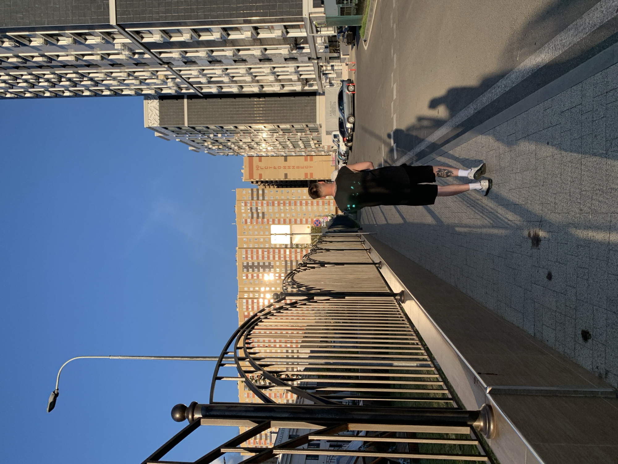 The image shows a young man from behind, standing on a sidewalk next to a road, with a large modern building in the background. He is wearing a black t-shirt and black shorts, and white socks with athletic shoes. He has a tattoo on his right calf. The sun is low in the sky, casting long shadows, suggesting it is either early morning or late afternoon. The sky is clear and blue, indicating good weather. The building behind him is a multi-story residential complex with many windows and balconies. The sidewalk is paved with square tiles. A black metal fence with a curved top runs along the edge of the sidewalk, separating it from what appears to be a lower level or embankment. Further in the background, more buildings are visible, some with brick facades. A car is parked on the road. The overall scene suggests an urban environment, likely a residential area. No discernible text is visible on the person or immediate surroundings, though there might be distant signage on the buildings not clear in the frame. The man's posture does not convey a strong emotion, appearing to be standing still, perhaps observing the surroundings.
