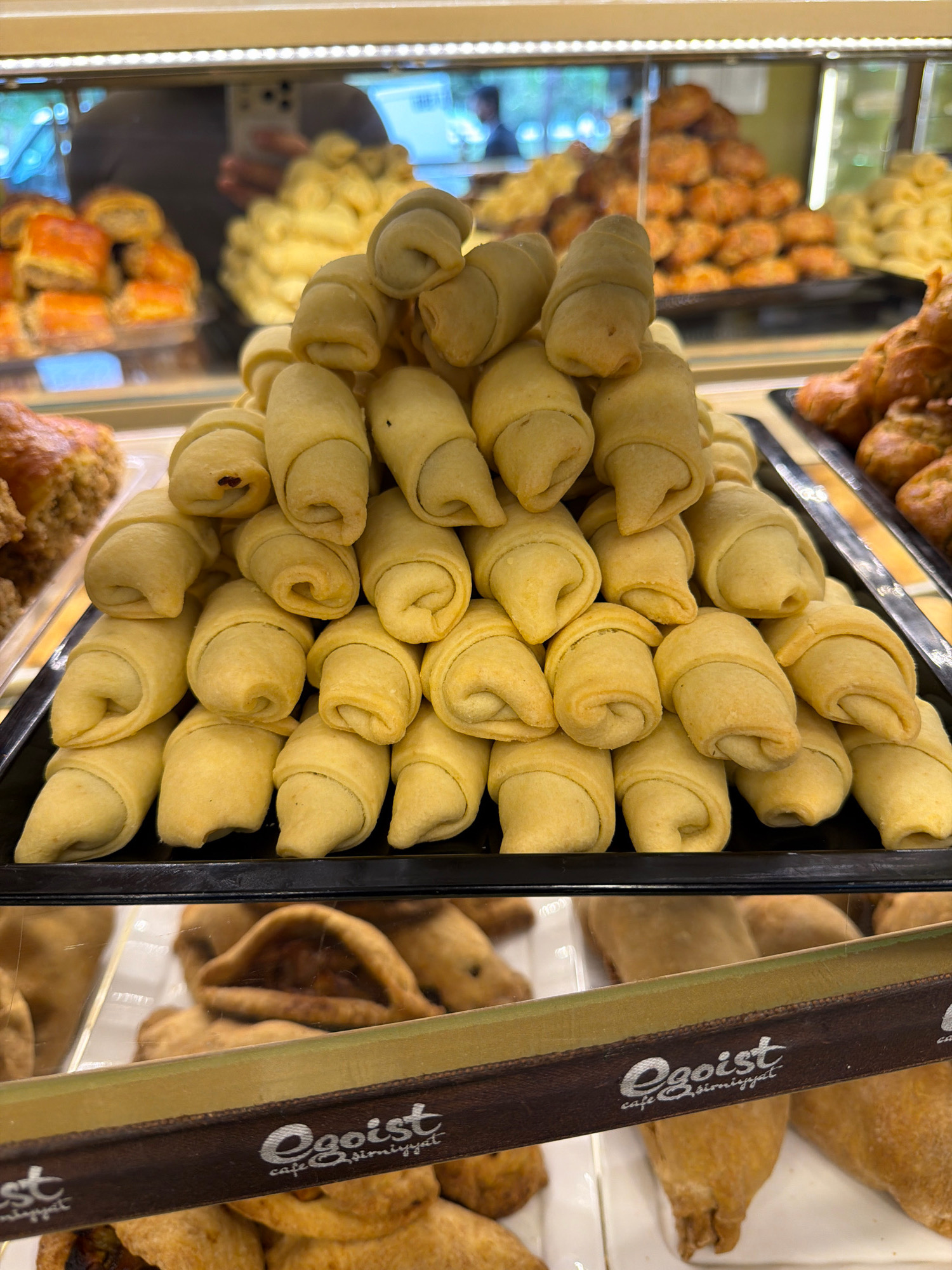 The image displays a close-up view of a bakery display case filled with various baked goods. In the foreground, a pyramid of crescent-shaped pastries, light golden in color and appearing to be made of a flaky dough, is prominently featured. These pastries are arranged in layers, creating a visually appealing stack.
Behind these pastries, a tray of what appears to be baklava or a similar layered dessert with a darker, syrupy glaze is visible. Further back, more pastries, possibly cookies or small cakes with a richer, browner hue, are stacked in trays, some appearing to be in a pyramidal formation. In the background, there are glimpses of what seem to be other bakery items, including mounds of light yellow, granular confectioneries and more trays of baked goods. The lighting suggests an indoor setting, likely a retail environment.
A brown band with white text is visible across the lower part of the image, containing the word "Egoist" and below it "cafe şirniyyat". This text indicates the name of the establishment, which is a cafe specializing in pastries or sweets (şirniyyat being Turkish/Azerbaijani for sweets/pastries). Given the presence of the word "şirniyyat", which is commonly used in Azerbaijan, and the general aesthetic of the bakery, it is highly probable that this establishment is located in Baku, Azerbaijan. The scene captures the abundance and variety of pastries offered in a typical bakery.