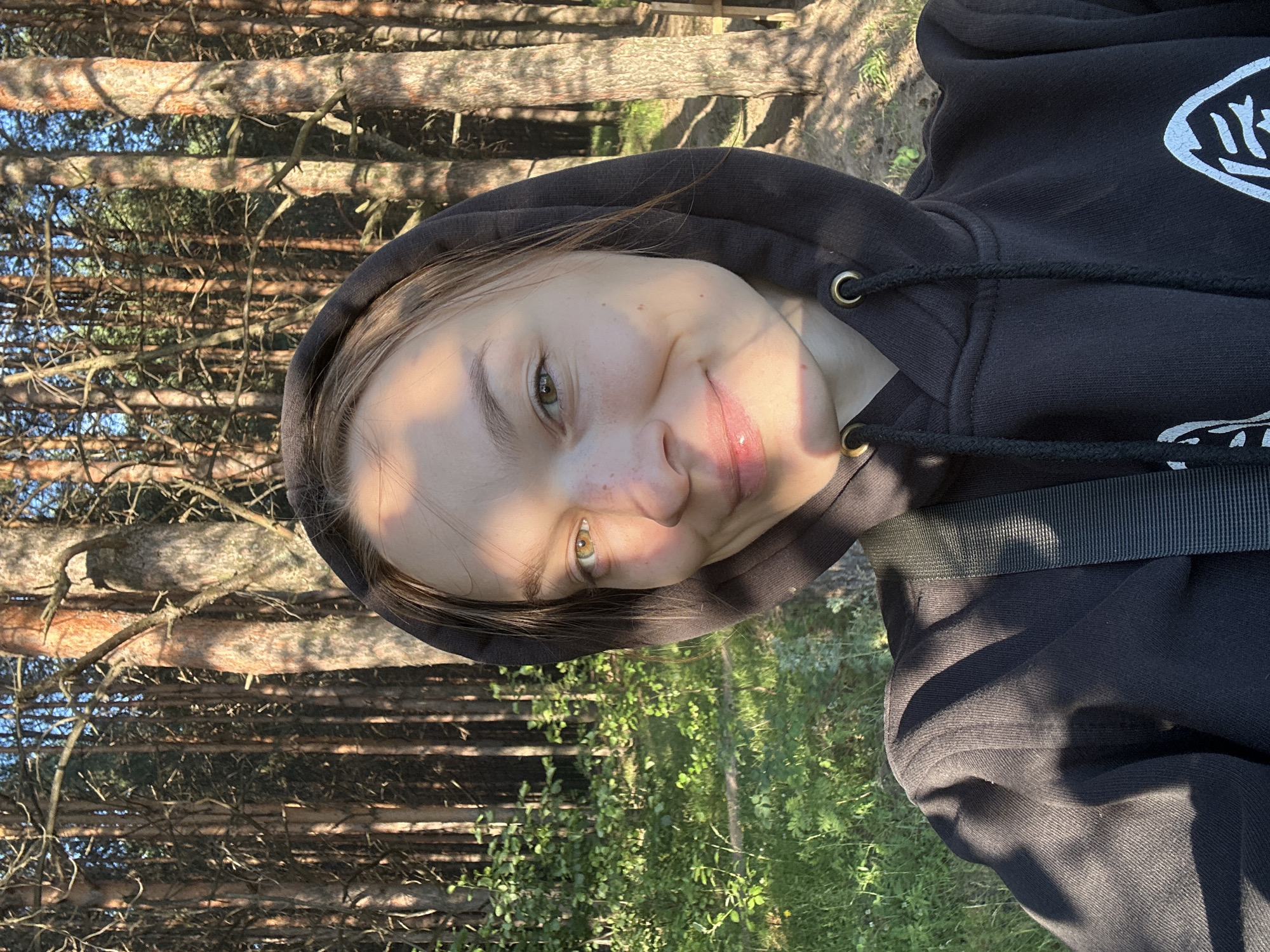 This is a portrait of a young person wearing a black hoodie. The person's face is turned towards the camera, with a slight smile. Their eyes are open and looking at the viewer. The background consists of a forest with tall pine trees and green undergrowth. The lighting suggests it is daytime, likely late afternoon due to the warm, dappled sunlight filtering through the trees. There are no visible objects other than the person and the natural environment. The overall mood appears relaxed and cheerful. The image was taken in Chaikovsky, Russia. No text is visible in the image.