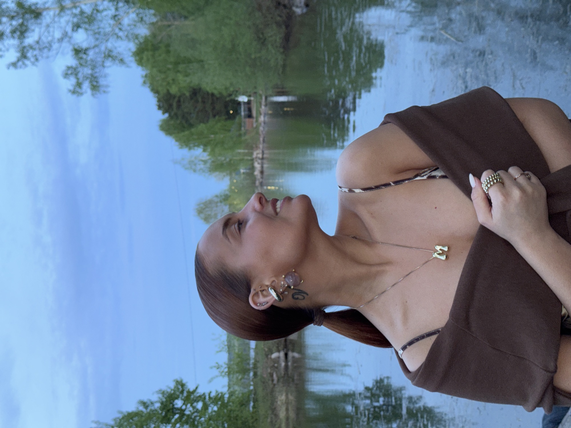 A woman is captured in a medium shot with a slightly upward angle, situated outdoors in what appears to be Chaikovsky, Russia. The background features a body of water, likely a pond or lake, with greenery visible along the shoreline. The sky is partly visible with some cloud cover. The woman is wearing a brown garment that drapes over her shoulders. She is also wearing a necklace with a letter "M" pendant. She has multiple rings on her hand. She is wearing an earring and a tattoo is visible. She is looking upwards and to the left.