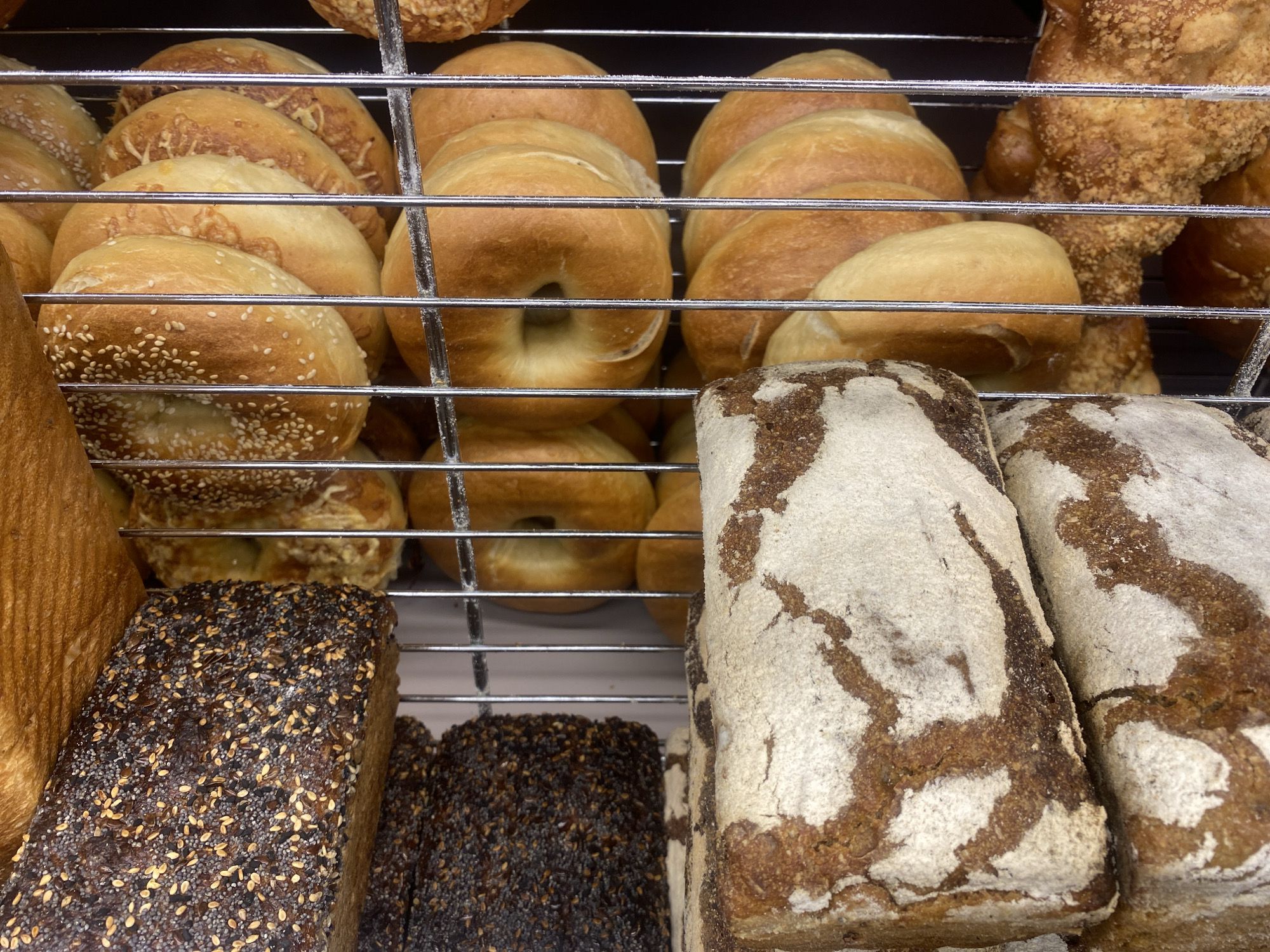 This is a medium-shot, eye-level photograph of a shelf filled with various types of bread. The subjects are primarily loaves of bread, presented in a supermarket or bakery setting. The bread types include round loaves with sesame seeds, larger round loaves, elongated loaves with dark crusts sprinkled with seeds, and loaves with a rustic, flour-dusted appearance. The bread is arranged on metal shelves, indicating a commercial display. There are no people visible in the frame, and the focus is solely on the baked goods. The lighting appears to be artificial, typical of an indoor retail environment. The overall scene is clean and organized, showcasing the bread as the main subject. There is no discernible text in the image. The location context is Warsaw, Poland, suggesting a Polish bakery or supermarket.