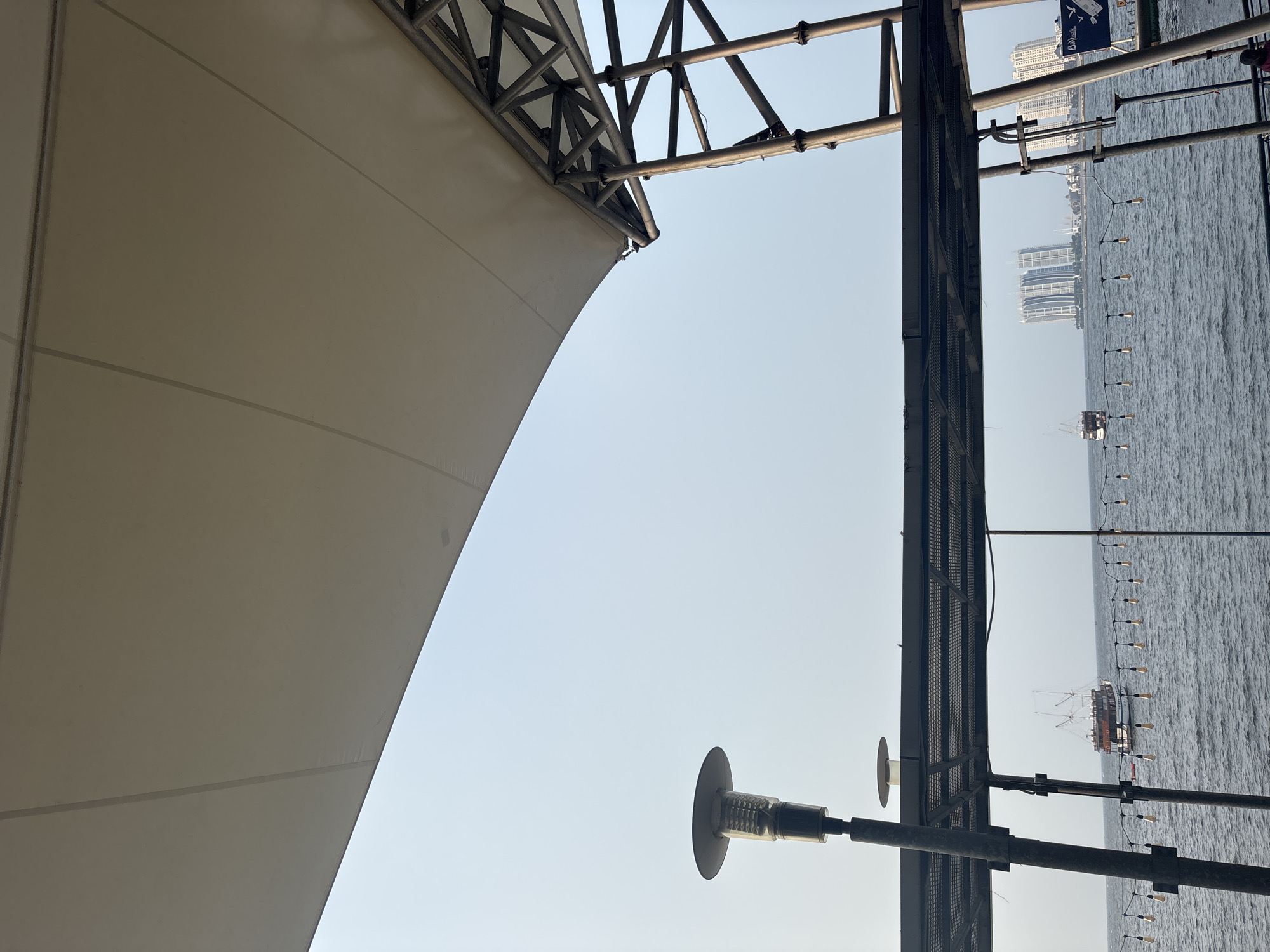 The image depicts a waterfront view in North Jakarta, Indonesia, likely from an elevated platform or balcony overlooking the sea. A large, light-colored canopy stretches across the upper portion of the frame, supported by a metallic structure. Below the canopy, a dark metal railing runs horizontally, and a lamppost with a modern design is visible on the left.
The sea occupies the middle and lower sections of the image, with gentle waves. Several vessels are scattered across the water: a larger boat with multiple masts is prominent in the middle left, and another smaller boat is closer to the right. A string of decorative lights hangs across the water, suspended by thin wires. In the background, a cityscape with several modern high-rise buildings is visible under a hazy, pale blue sky. The overall scene suggests a relaxed outdoor setting, possibly a restaurant or recreational area, in a coastal urban environment.
