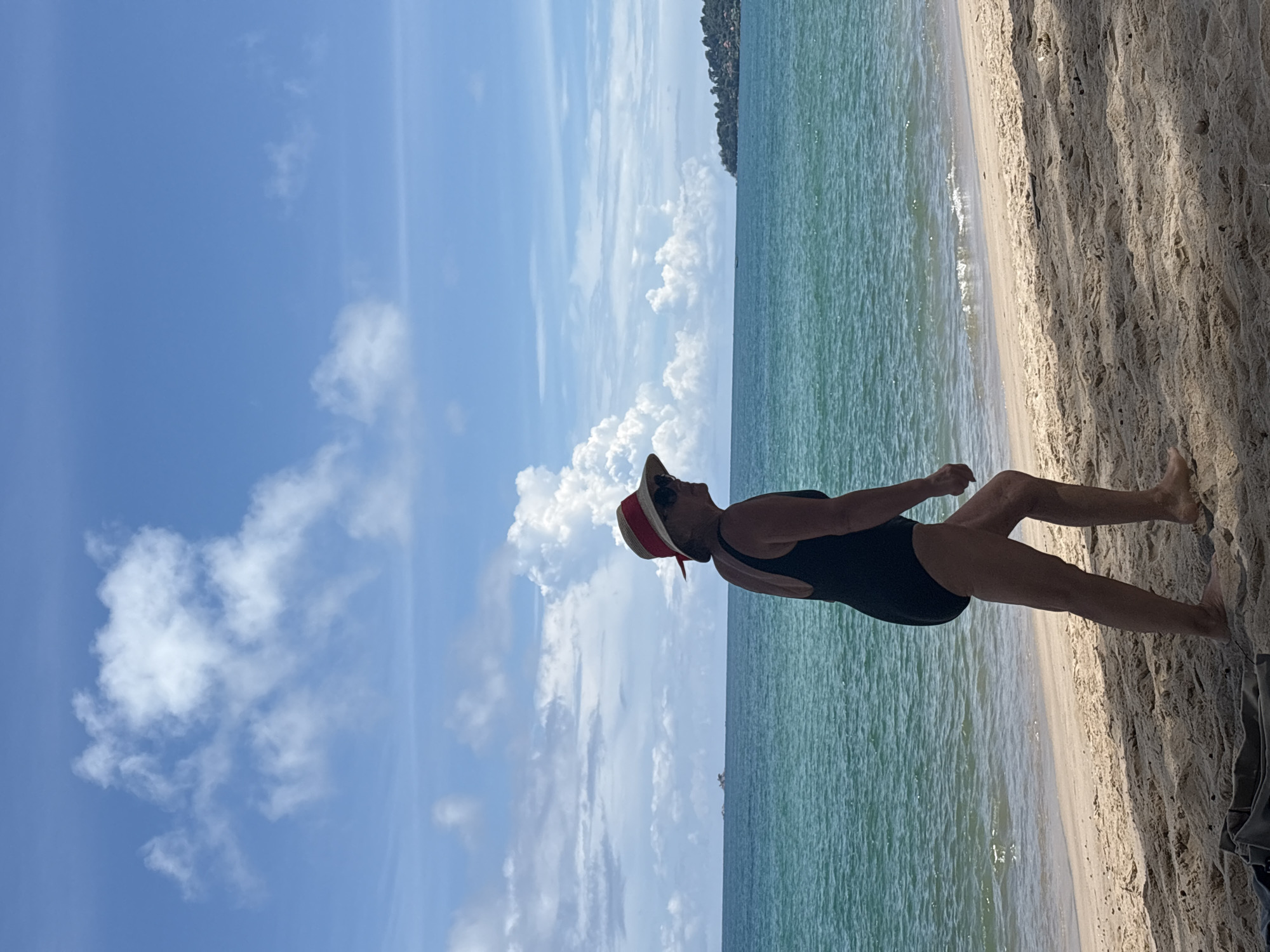 An adult female wearing a black one-piece swimsuit and a straw hat with a red ribbon is walking along a sandy beach. The ocean, with its turquoise water, is to her left, and the beach continues to her right. In the distance, a small boat is visible on the water, and a treed peninsula is on the horizon. The sky is blue with scattered white clouds. The scene appears to be in Choeng Thale, Thailand.