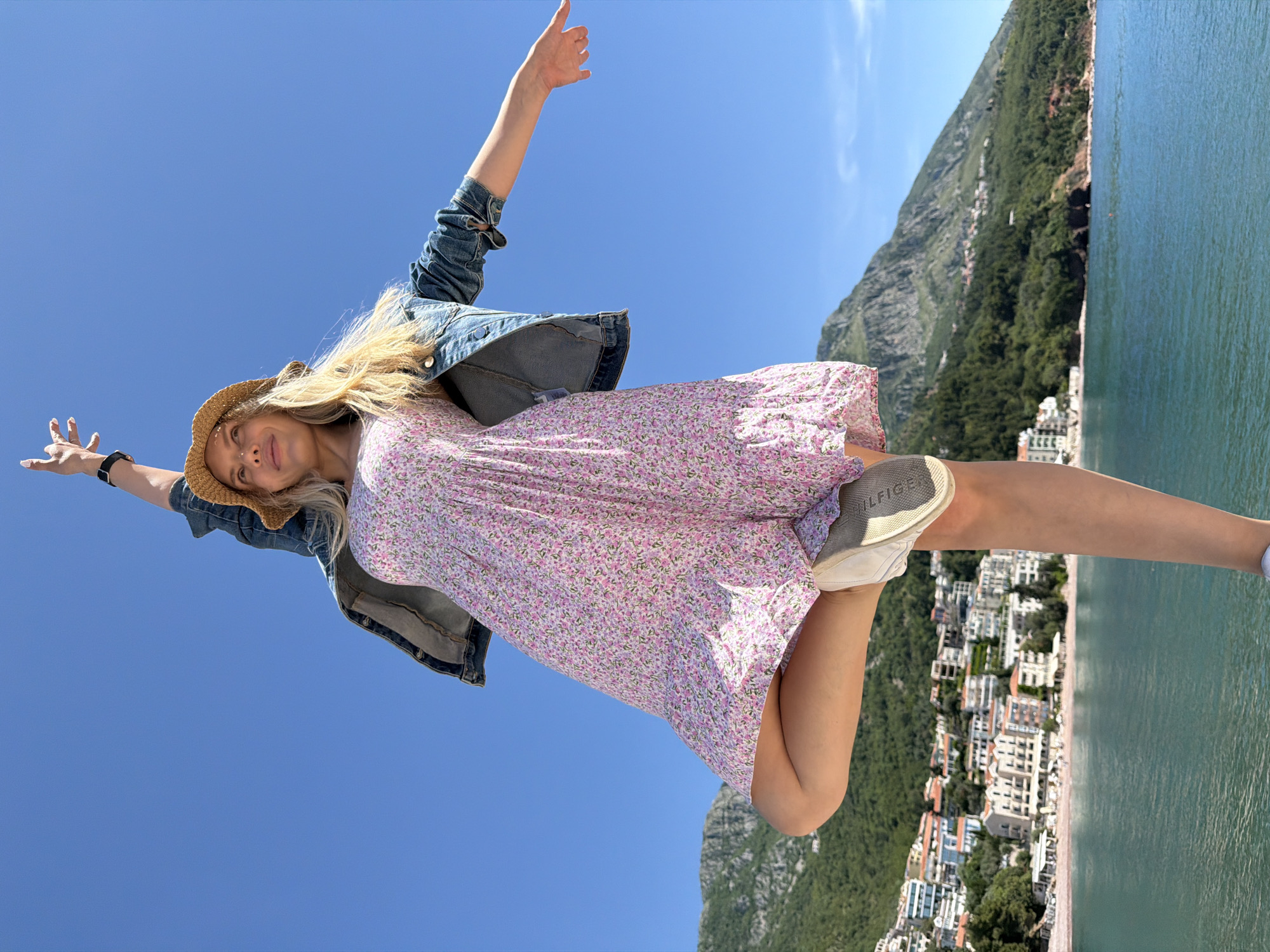 A woman in a straw hat, denim jacket, and pink floral dress is seen in a dynamic pose with one leg extended towards the camera, captured from a low angle. The background shows the coastline of Sveti Stefan, Montenegro, featuring mountains, buildings, and the Adriatic Sea. The woman's arms are outstretched, suggesting a joyful or celebratory gesture. The sky is clear blue, indicating a sunny day.