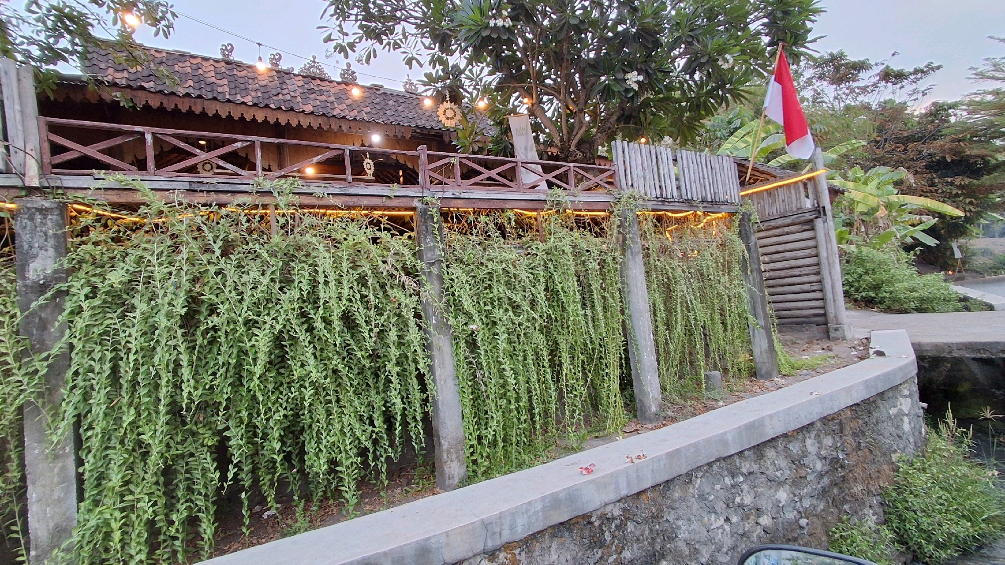 The image shows an outdoor scene at dusk, likely a restaurant or cafe in Banguntapan, Indonesia. A two-story wooden structure with a tiled roof is visible, adorned with string lights and ornate trim. A red and white flag, likely the Indonesian flag, flies from a pole on the right side of the structure. The lower level of the structure is obscured by a dense growth of cascading green plants hanging from a railing. A wooden staircase leads up to the second level on the right. In the foreground, a concrete wall with a stone base runs along a pathway. The environment appears lush and tropical, with trees and foliage visible in the background. No people are present in the frame.