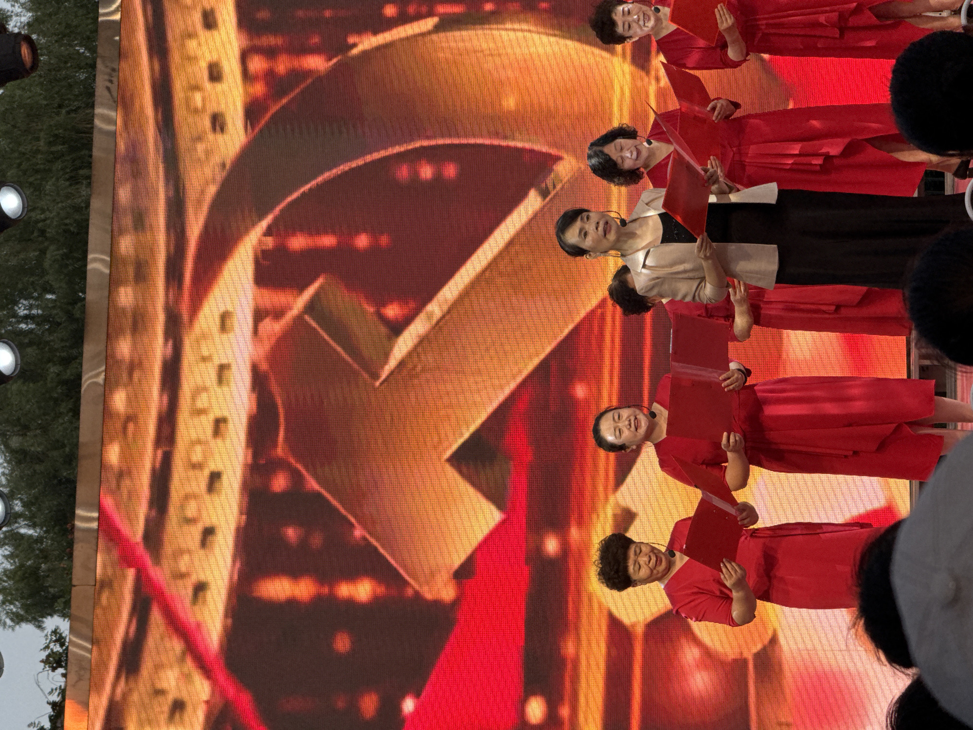 A group of six women, dressed in red dresses and one woman in a beige jacket over a black dress, stand on a stage in front of a large screen displaying a golden hammer and sickle emblem. The women are holding red folders and appear to be singing or reciting. They are all facing forward, towards an unseen audience. The screen behind them is illuminated with red and gold light. The scene appears to be an outdoor event, with greenery visible at the top of the image. The presence of the hammer and sickle emblem suggests a political or patriotic event, possibly related to the Communist Party of China. The context of Jinzhou, China is absent from the visual information provided by the image.