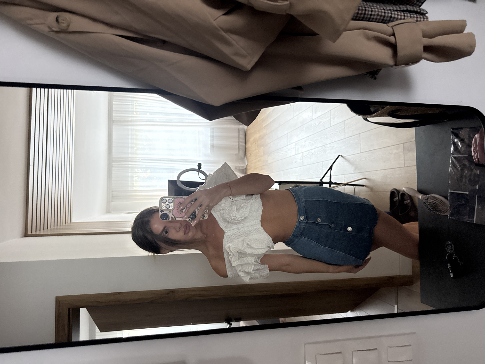 A young woman with dark hair pulled back stands in front of a full-length mirror, taking a selfie with her phone. She is wearing an off-the-shoulder white ruffled crop top and a denim mini-skirt with a button-down front. Her reflection shows a doorway with a wooden door and frame to her left, and a brightly lit room with sheer curtains in the background. To her right, the mirror reflects a beige trench coat hanging on a hook and part of a checkered garment. The floor in the background is light wood. On the dark surface in front of the mirror are keys, a hairbrush, and what appears to be a plastic bag containing clothing. The scene is indoors, in what appears to be a residential setting. There is no indication of the city of Plovdiv, Bulgaria.
