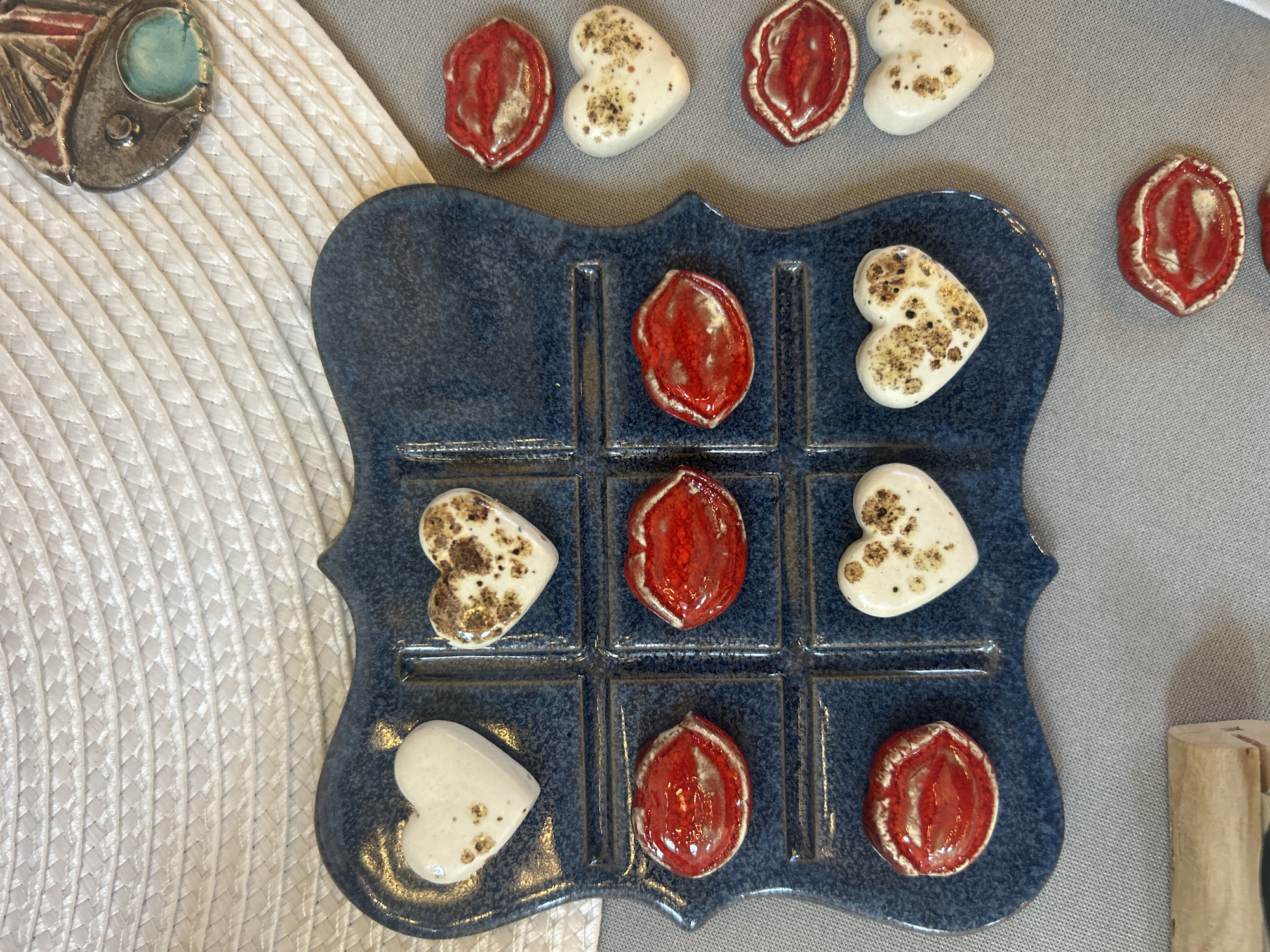 The image depicts a handmade tic-tac-toe board game set on a neutral-colored surface. The board itself is blue with a glossy, possibly ceramic finish, and the grid is defined by raised lines. The game pieces consist of two types: heart-shaped pieces with a white base and brown speckles, and oval-shaped pieces that are red with a textured surface. The board is partially filled with the game pieces, and some pieces of both types are scattered around the board on the surrounding surface. The scene is lit with soft, diffused light. To the left of the board is a white textured mat and a ceramic object of blue and silver color. To the right of the board is a light-colored cylindrical object. There is no information indicating this image was taken in the city of Sofia, Bulgaria.