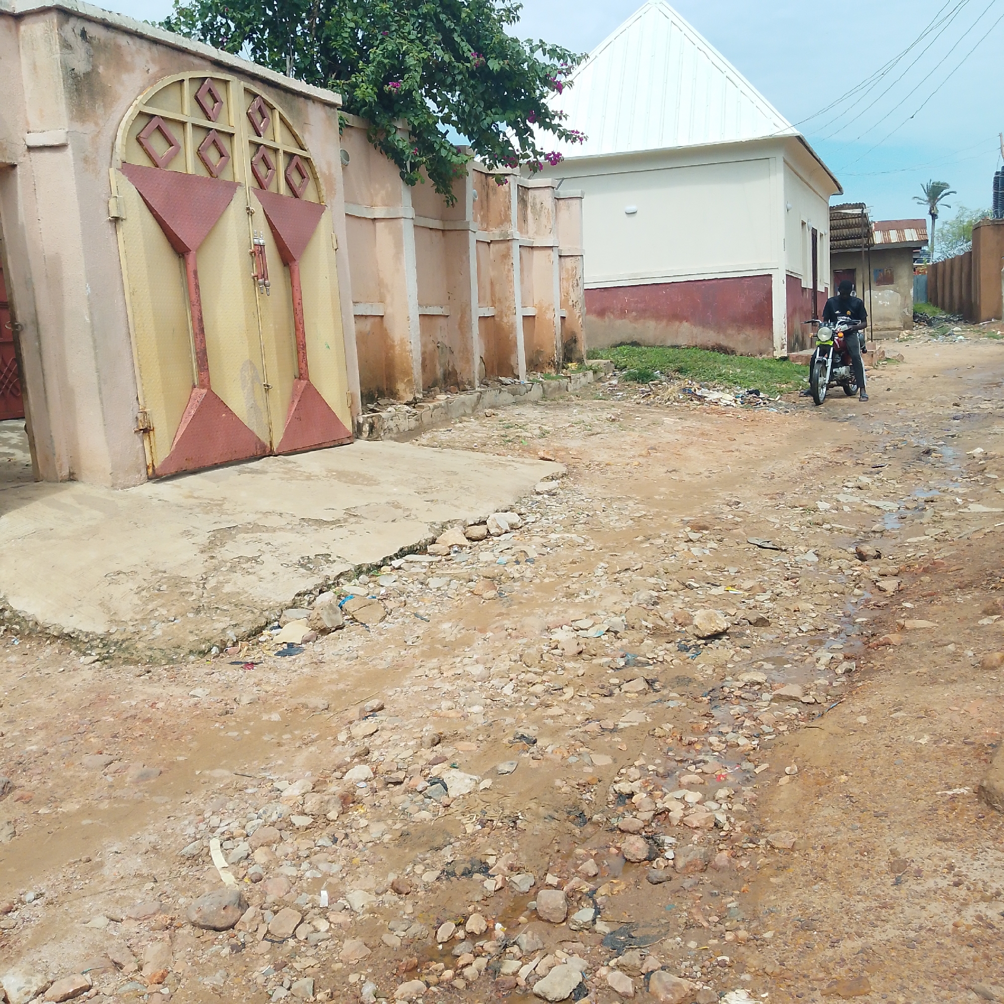 A person rides a motorcycle on a dirt road in an urban area. The person is wearing a black shirt and pants and a black mask. The motorcycle is red. The road is unpaved and covered with rocks and debris. There are buildings on either side of the road. One building has a large yellow and red metal gate. Another building has a white roof. There are trees and bushes in the background. The sky is cloudy. The image appears to have been taken in the daytime.