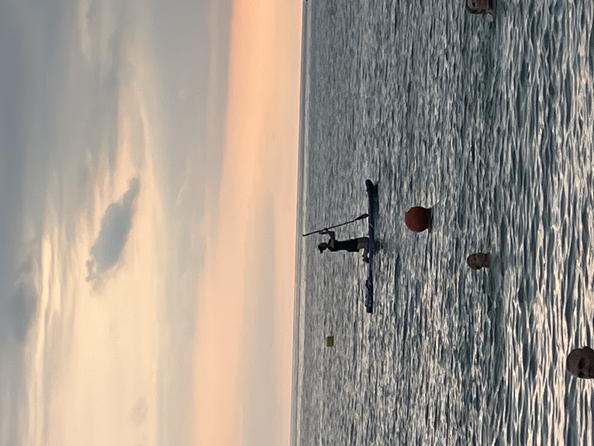 This image captures a serene seascape at sunset, likely near the city of Sochi, Russia. The sky is a gradient of soft orange and pink hues, with wispy clouds scattered across the horizon. The water is calm, reflecting the sky's colors and creating gentle ripples.
In the middle ground, a person is paddleboarding. They are kneeling on the board, holding a paddle and facing towards the left of the frame. The paddleboarder is silhouetted against the lighter sky. A yellow buoy is visible to the left of the paddleboarder.
In the foreground, several people are partially submerged in the water, with their heads and shoulders visible. They appear to be swimming or floating. One person is near the center of the frame, looking towards the left. Another person is further to the right, also in the water. A large orange buoy is situated between the paddleboarder and the foreground swimmers. In the bottom left corner, a man's head is visible, looking towards the right.