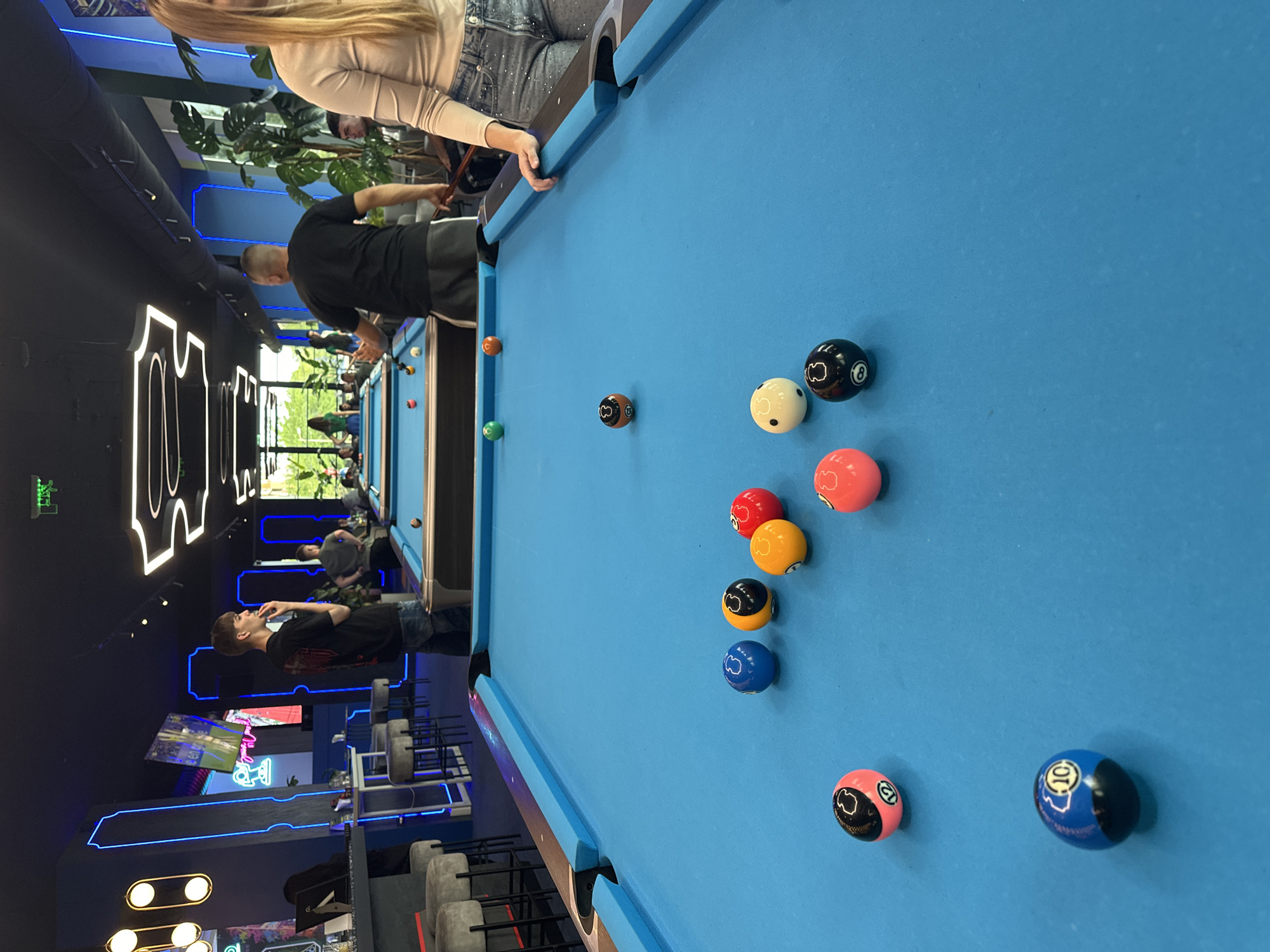 The image depicts a pool hall in Varna, Bulgaria. A bright blue pool table dominates the foreground, displaying a set of pool balls scattered across its surface. A woman's hand rests on the edge of the table near the top of the frame, while two people stand in the background near the table. Further back, multiple pool tables are visible, occupied by other people. The interior has a contemporary design, with neon lights illuminating the walls and ceilings. High-top chairs and tables are arranged to the left of the pool tables, and a television screen is visible on the wall. Through the windows, the scenery of the city is visible.