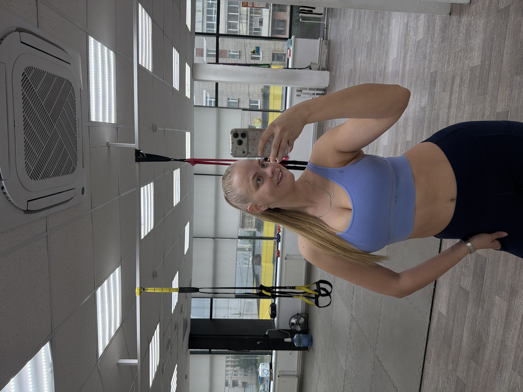 A woman is taking a selfie in a gym in what appears to be Izhevsk, Russia. She is wearing a blue sports bra and dark leggings. The gym has large windows that look out onto a city street. There are suspension training straps hanging from the ceiling, as well as other gym equipment visible in the background. The ceiling is tiled with recessed lighting and a large air conditioning unit. The floor is covered in a light-colored wood-patterned floor.
