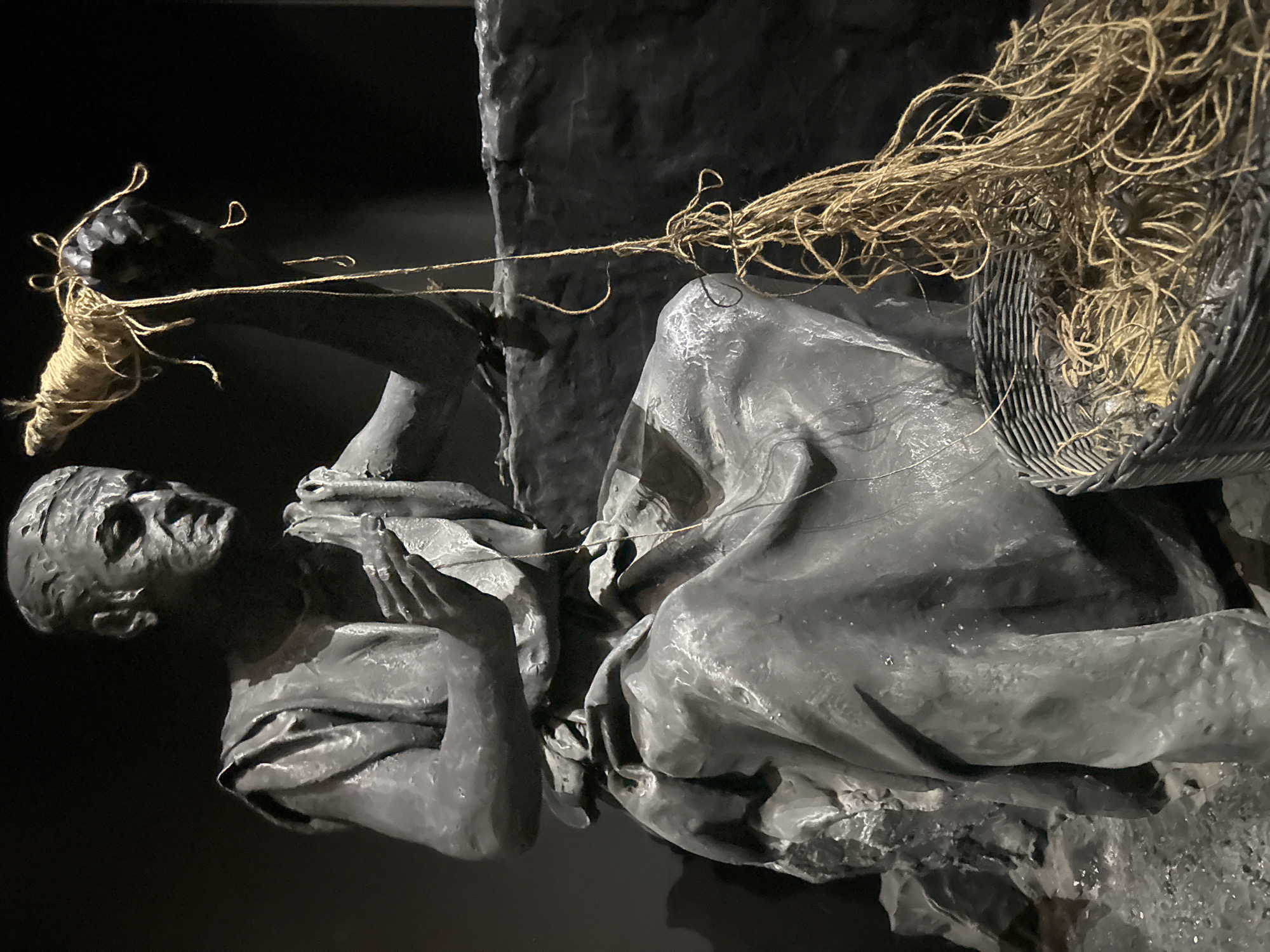 A full-body statue of a person is depicted, seated and engaged in the act of spinning yarn. The statue, rendered in a dark, textured material, portrays a figure with an aged face and draped clothing, characteristic of ancient attire. The individual's right hand holds a dark, rough object around which light-colored yarn is wound, while the left hand appears to be in the process of spinning. A basket filled with more yarn sits at the figure's feet. The background is dark and indistinct, creating a spotlight effect on the statue. The statue is located in the Tamar Regional Council, Israel.
