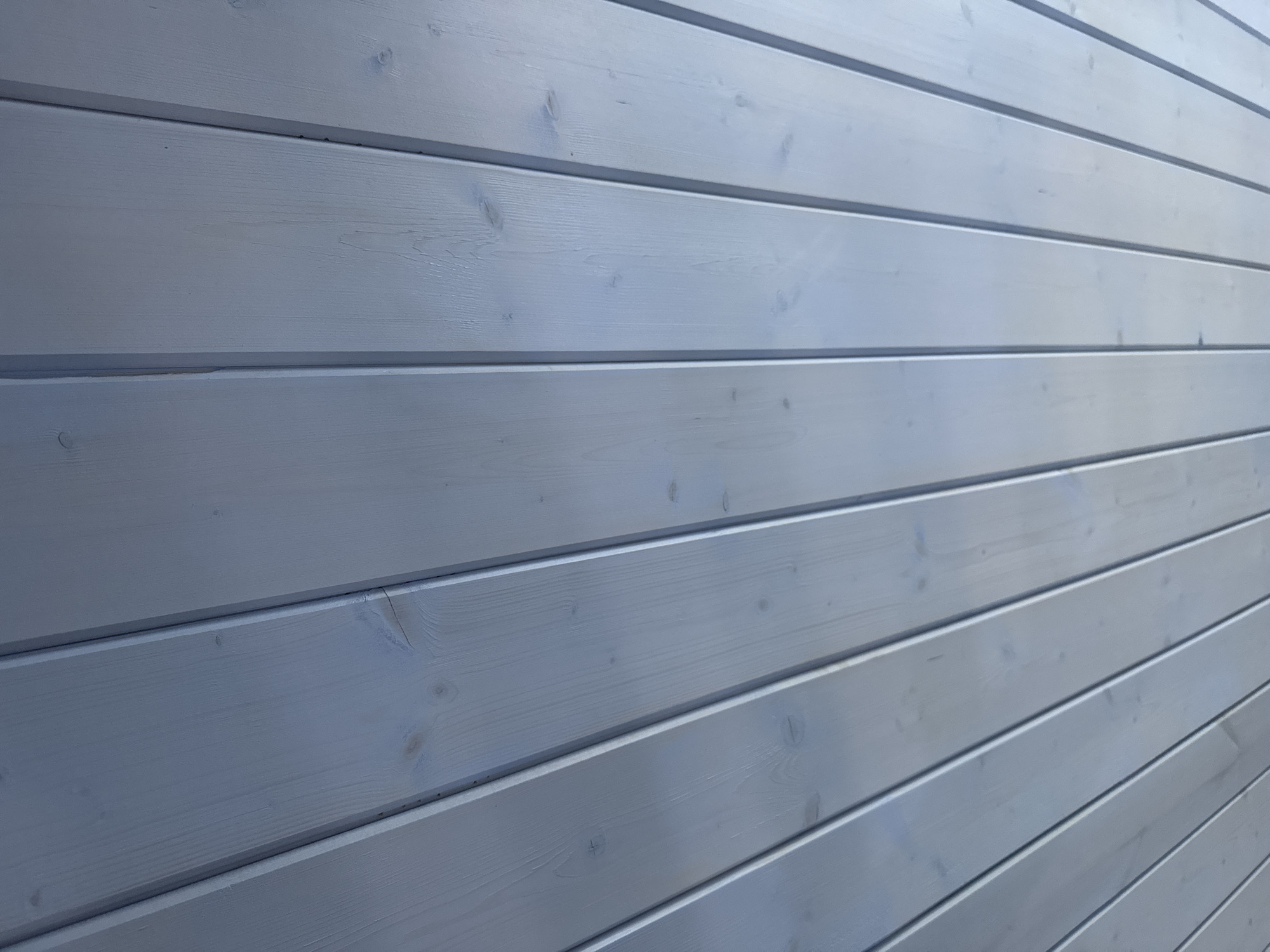 The image shows a close-up, angled view of a light blue painted wooden ceiling composed of parallel planks. The planks are positioned diagonally from the bottom left to the top right of the frame. There are visible wood grain patterns and some knot details on the planks, indicating the material is natural wood. The paint appears to be a light, semi-transparent wash, allowing some of the wood texture to show through. The lines between the planks are distinct, creating a striped pattern. The lighting suggests an ambient, possibly overcast or indoor light source, casting subtle shadows in the grooves between the planks. The location is specified as Doma 1096 Km, Russia. No people or specific objects other than the ceiling itself are visible in the frame. The scene is static and devoid of action.