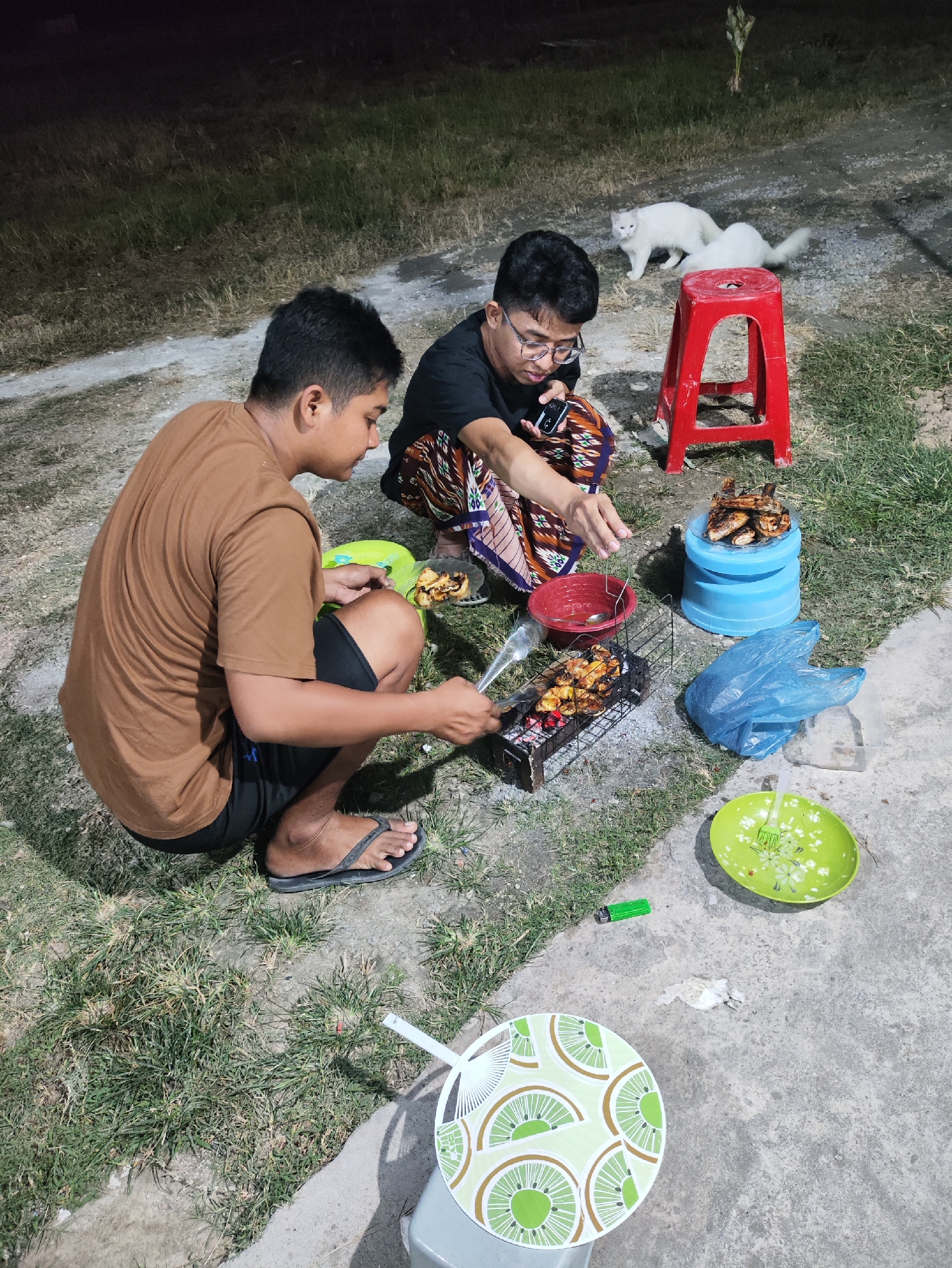 The image depicts a nighttime scene outdoors, likely in a residential or common area in Prague, Indonesia, given the presence of grass and concrete surfaces. Two young males are crouched on the ground, engaged in grilling food. One individual, wearing a brown t-shirt and black shorts, is using tongs to handle food on a charcoal grill. The other, wearing a black t-shirt and patterned pants, appears to be preparing food or observing the grilling process, holding a phone in one hand and gesturing with the other.
Several objects are scattered around them, including a red stool, a blue plastic container with grilled food on top, a red bowl with a spoon, a green plastic bag, a green plate with a fork, a lighter, and a round serving tray with a kiwi pattern. Two white cats are visible in the background, one standing and another partially obscured. The ground is a mix of grass and concrete, with some areas appearing dimly lit by an unseen light source. The overall atmosphere suggests a casual outdoor meal or gathering.