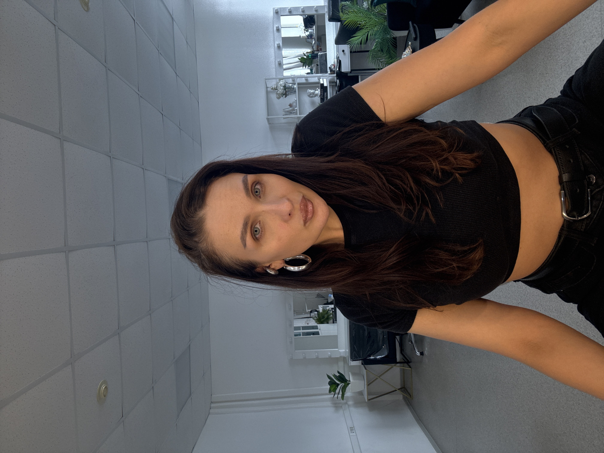 A woman with long, dark brown hair and light green eyes poses for a selfie in what appears to be a studio or salon. She is wearing a black cropped top and black jeans with a belt. The location is likely indoors, given the ceiling tiles and fluorescent lighting. In the background, there is a vanity mirror with lights, shelves with decorative items, and a green plant. The image is taken at an angle, with the camera pointing slightly downward. The city and country of Chaikovsky, Russia are unable to be confirmed by visual data alone.