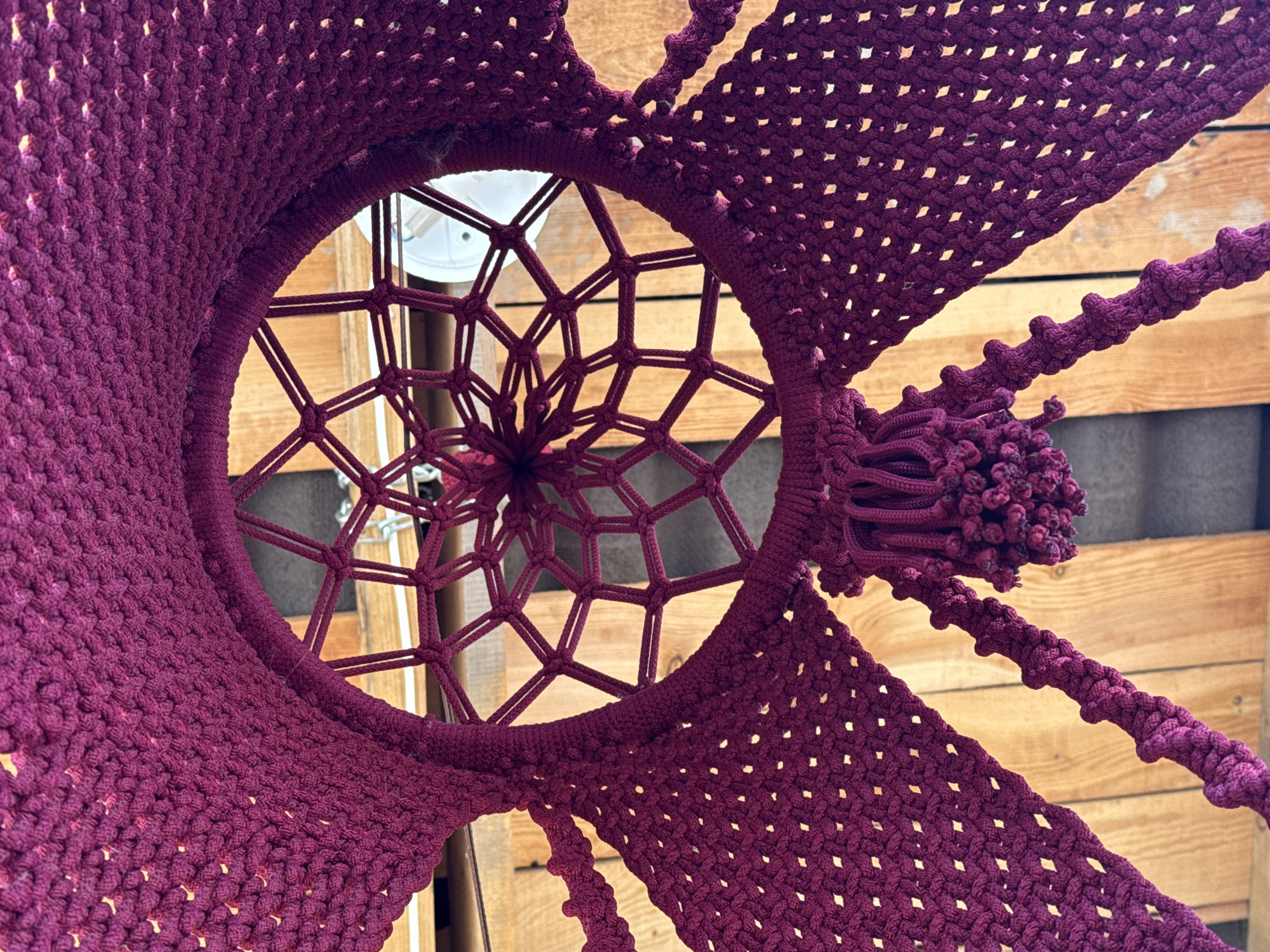 The image shows a maroon macrame hanging chair from a low-angle perspective, looking up towards a light fixture on the ceiling. The chair's woven structure forms a circular opening at the top, revealing the white light fixture and portions of a wooden slat wall behind it. The macrame is intricately knotted, forming a dense, textured pattern throughout the body of the chair, with braided ropes ending in tassel-like knots hanging down. The location is indeterminable without additional context.