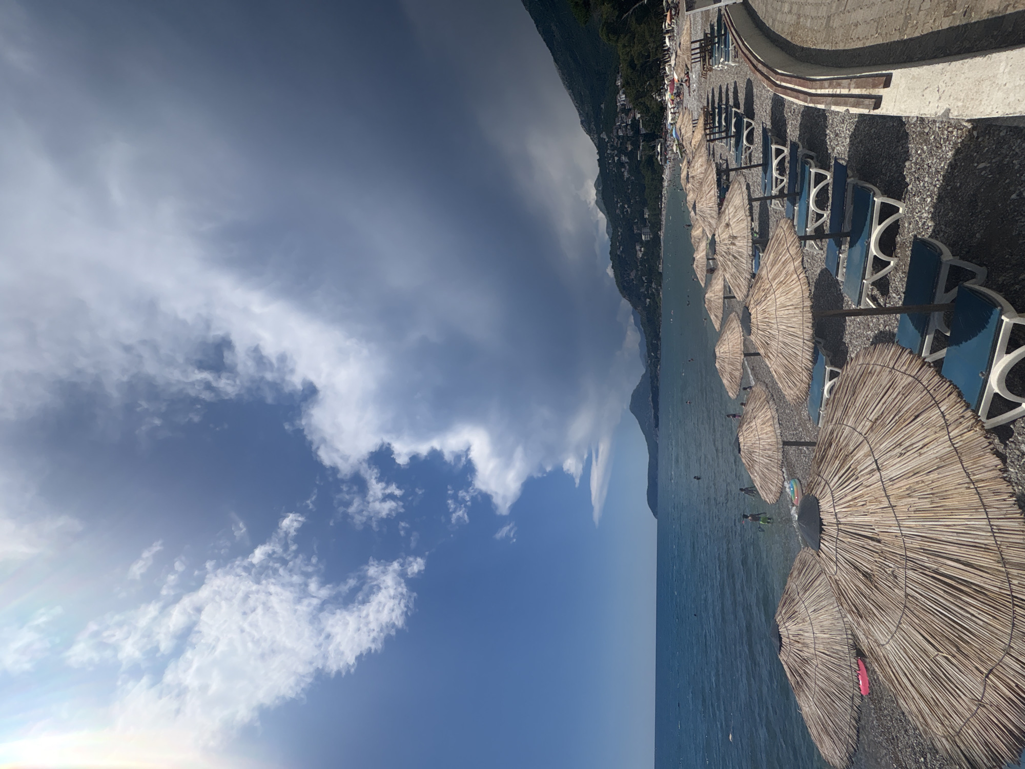 This is a landscape-oriented photograph taken at a beach in Bar, Montenegro. The scene depicts a shoreline lined with beach chairs and straw umbrellas, facing a calm, blue body of water. In the background, a lush green hillside rises steeply, dotted with buildings, suggesting a town or village built into the mountainside. The sky is a dramatic mix of deep blue with large, wispy clouds, indicating a day with varied weather, possibly transitioning between sun and shade.
The beach itself is covered in dark pebbles. Numerous blue and white beach chairs are neatly arranged under the straw umbrellas, ready for sunbathers. Some people are visible in the shallow water, swimming or wading. The overall atmosphere appears to be one of a relaxed summer day at a popular coastal destination. The sun is casting shadows, suggesting it is likely mid-day or early afternoon. There is no visible text in the image.